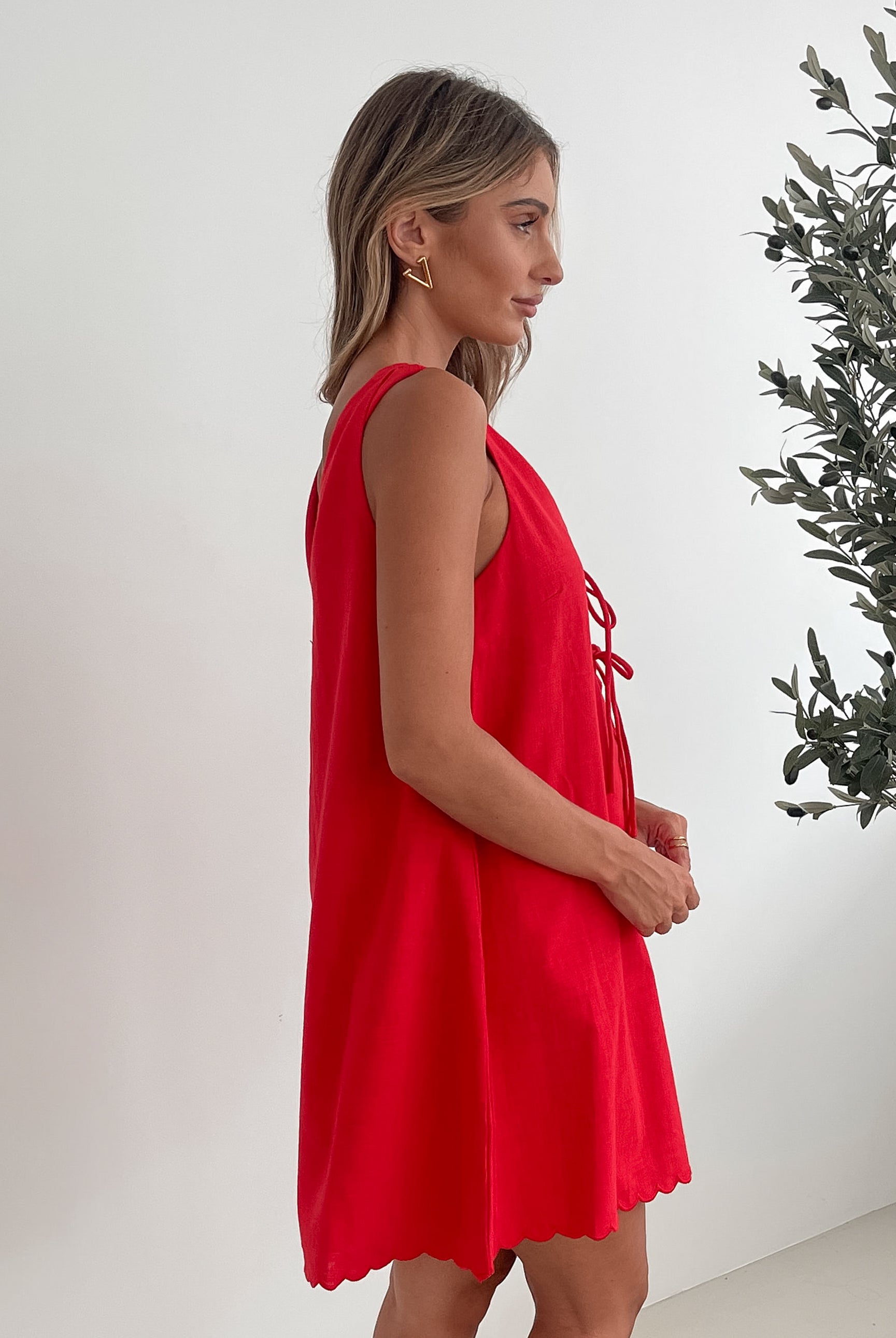 A woman with long, wavy blonde hair stands indoors in front of a plain white wall, with a green leafy plant beside her. She is wearing a sleeveless red dress with a V-neck and two front tie details, similar in style to the previous dress, featuring a scalloped hem. Her hands are gently clasped together in front of her, and she has a subtle smile on her face. She accessorizes with gold hoop earrings and a delicate ring, and her makeup is natural and polished.