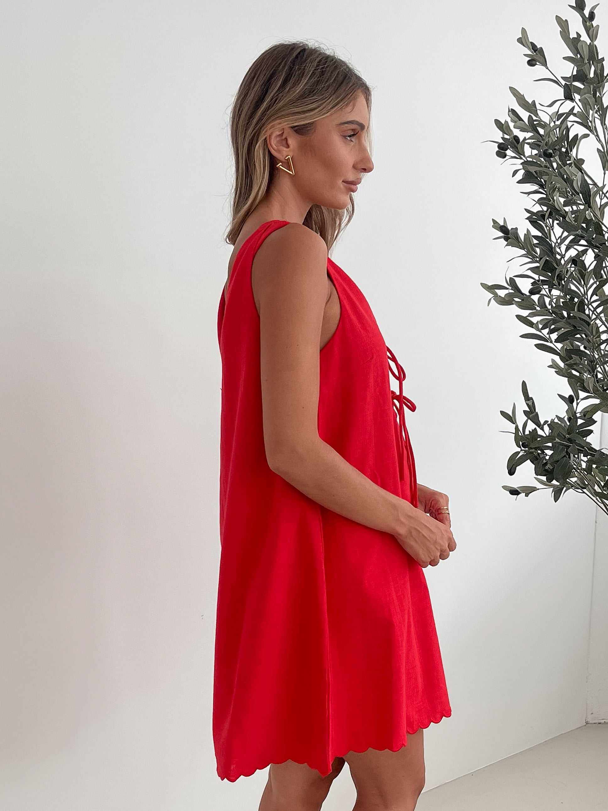 A woman with long, wavy blonde hair stands indoors in front of a plain white wall, with a green leafy plant beside her. She is wearing a sleeveless red dress with a V-neck and two front tie details, similar in style to the previous dress, featuring a scalloped hem. Her hands are gently clasped together in front of her, and she has a subtle smile on her face. She accessorizes with gold hoop earrings and a delicate ring, and her makeup is natural and polished.