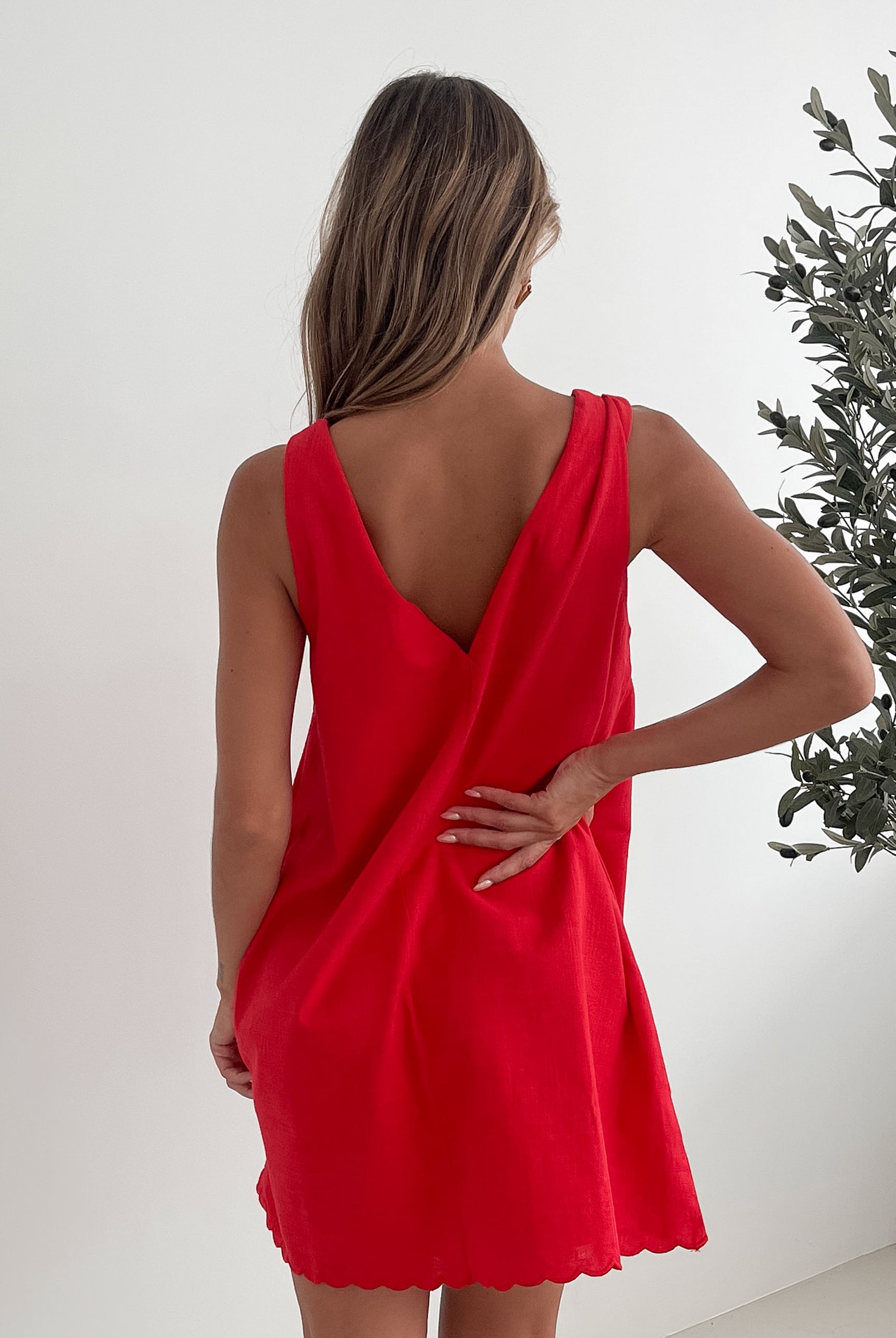 A woman with long, wavy blonde hair stands indoors in front of a plain white wall, with a green leafy plant beside her. She is wearing a sleeveless red dress with a V-neck and two front tie details, similar in style to the previous dress, featuring a scalloped hem. Her hands are gently clasped together in front of her, and she has a subtle smile on her face. She accessorizes with gold hoop earrings and a delicate ring, and her makeup is natural and polished.