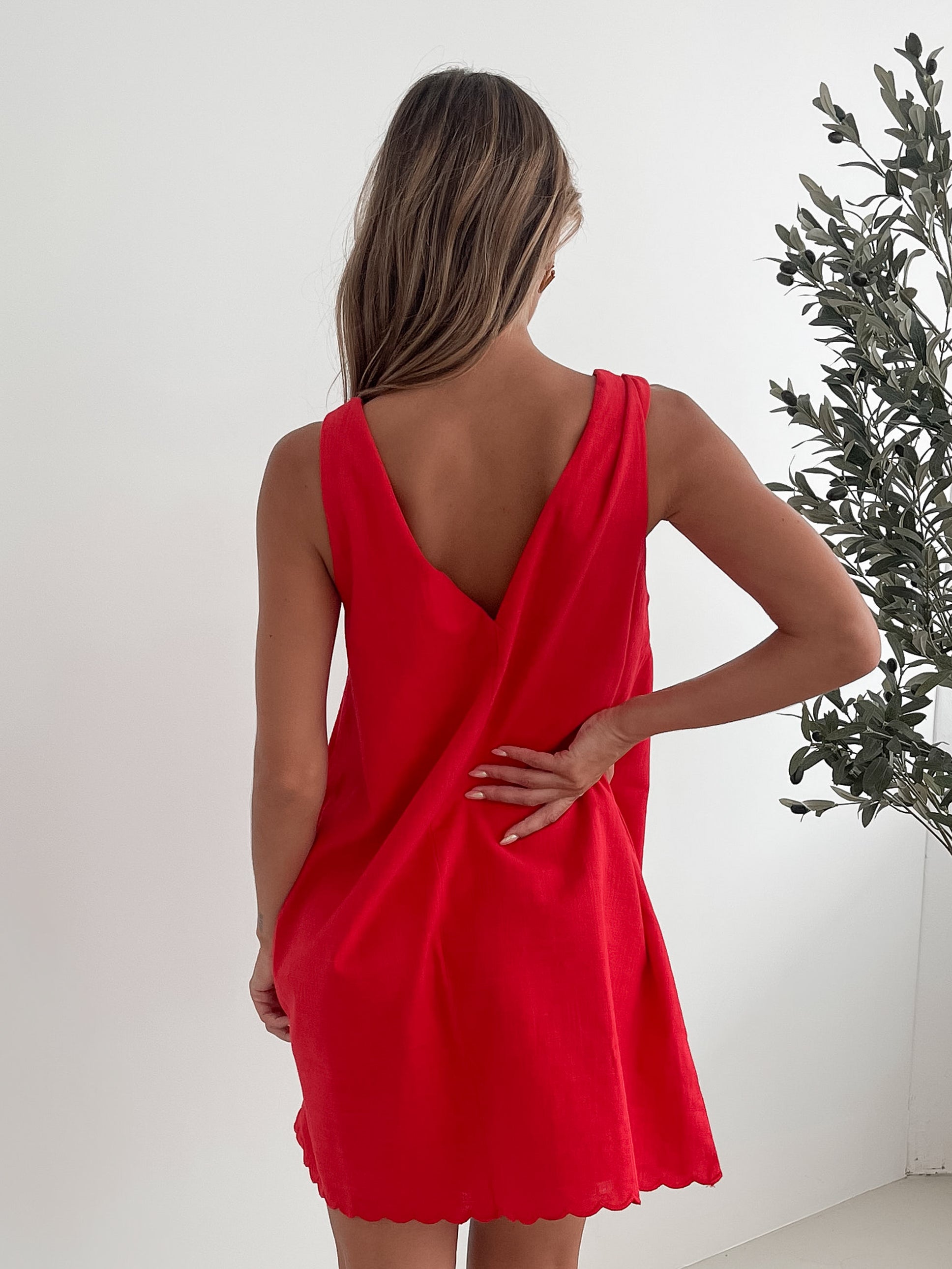 A woman with long, wavy blonde hair stands indoors in front of a plain white wall, with a green leafy plant beside her. She is wearing a sleeveless red dress with a V-neck and two front tie details, similar in style to the previous dress, featuring a scalloped hem. Her hands are gently clasped together in front of her, and she has a subtle smile on her face. She accessorizes with gold hoop earrings and a delicate ring, and her makeup is natural and polished.