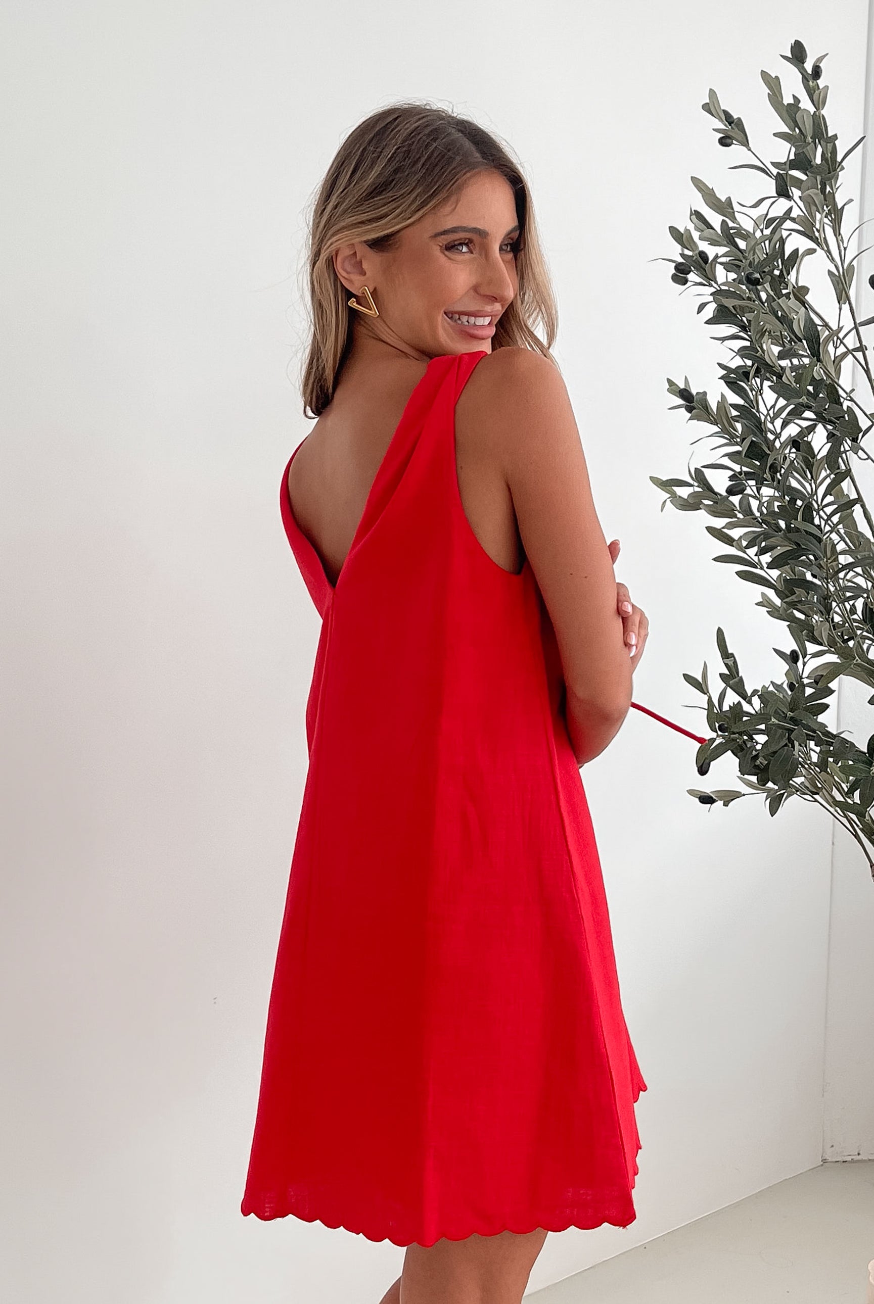 A woman with long, wavy blonde hair stands indoors in front of a plain white wall, with a green leafy plant beside her. She is wearing a sleeveless red dress with a V-neck and two front tie details, similar in style to the previous dress, featuring a scalloped hem. Her hands are gently clasped together in front of her, and she has a subtle smile on her face. She accessorizes with gold hoop earrings and a delicate ring, and her makeup is natural and polished.