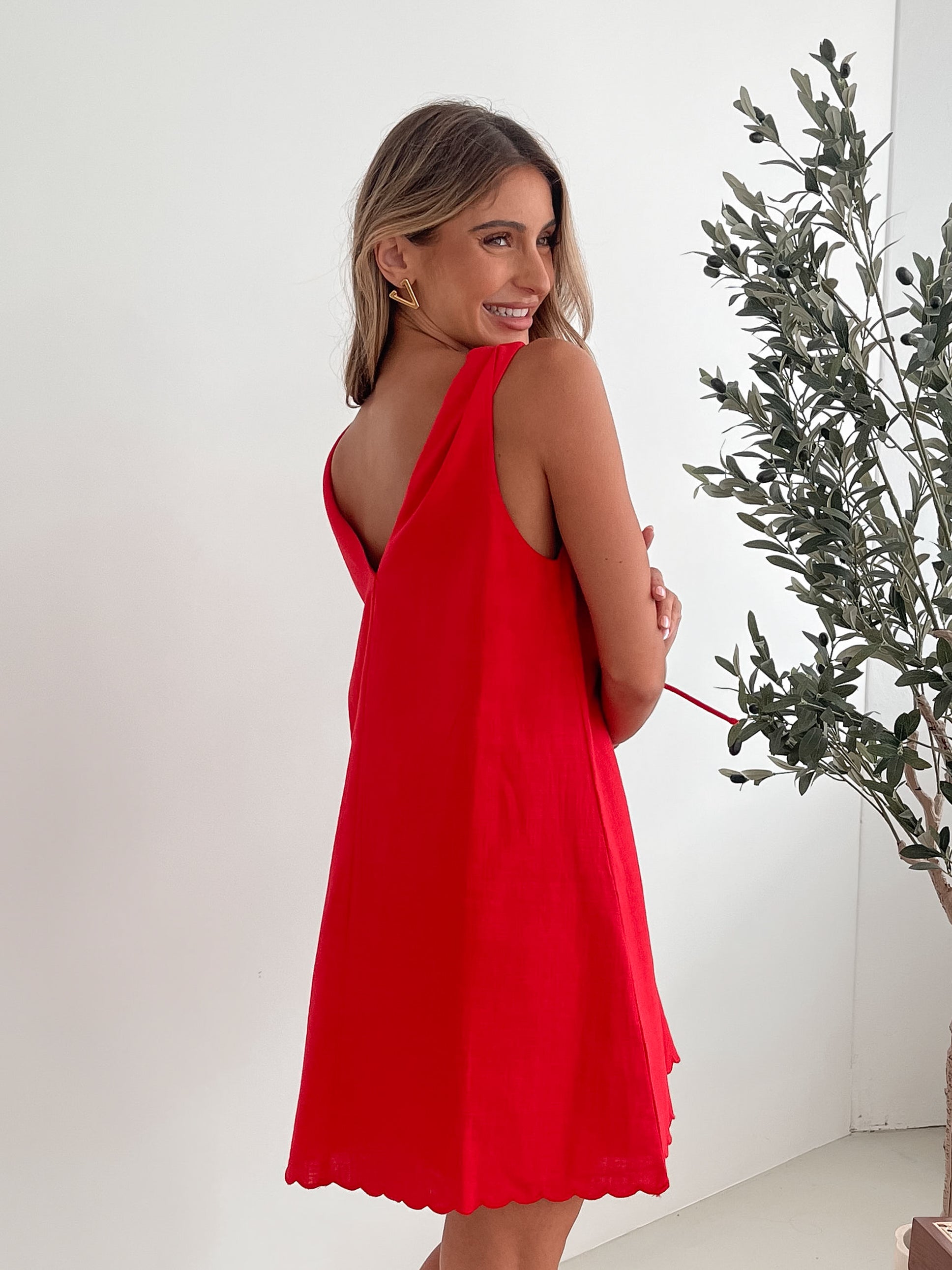 A woman with long, wavy blonde hair stands indoors in front of a plain white wall, with a green leafy plant beside her. She is wearing a sleeveless red dress with a V-neck and two front tie details, similar in style to the previous dress, featuring a scalloped hem. Her hands are gently clasped together in front of her, and she has a subtle smile on her face. She accessorizes with gold hoop earrings and a delicate ring, and her makeup is natural and polished.