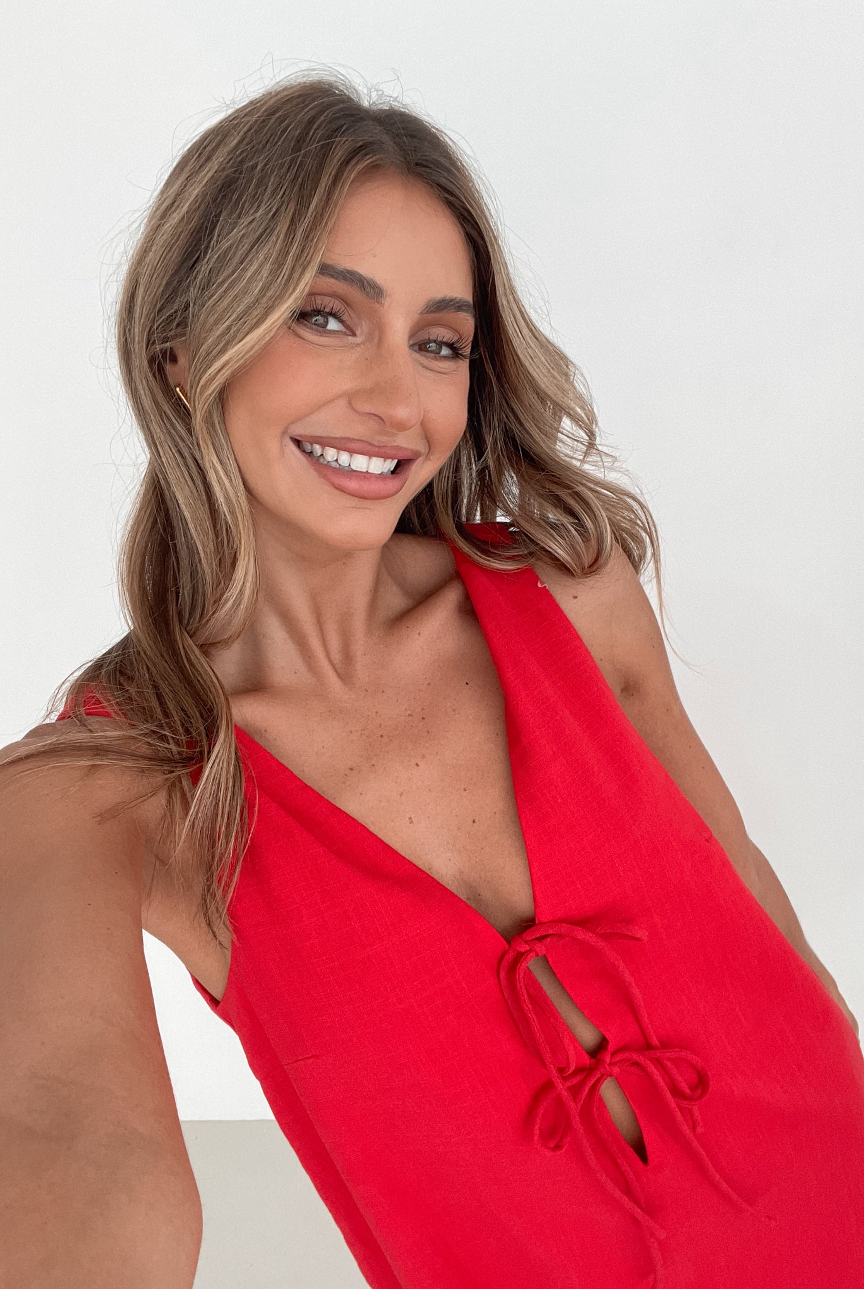 A woman with long, wavy blonde hair stands indoors in front of a plain white wall, with a green leafy plant beside her. She is wearing a sleeveless red dress with a V-neck and two front tie details, similar in style to the previous dress, featuring a scalloped hem. Her hands are gently clasped together in front of her, and she has a subtle smile on her face. She accessorizes with gold hoop earrings and a delicate ring, and her makeup is natural and polished.