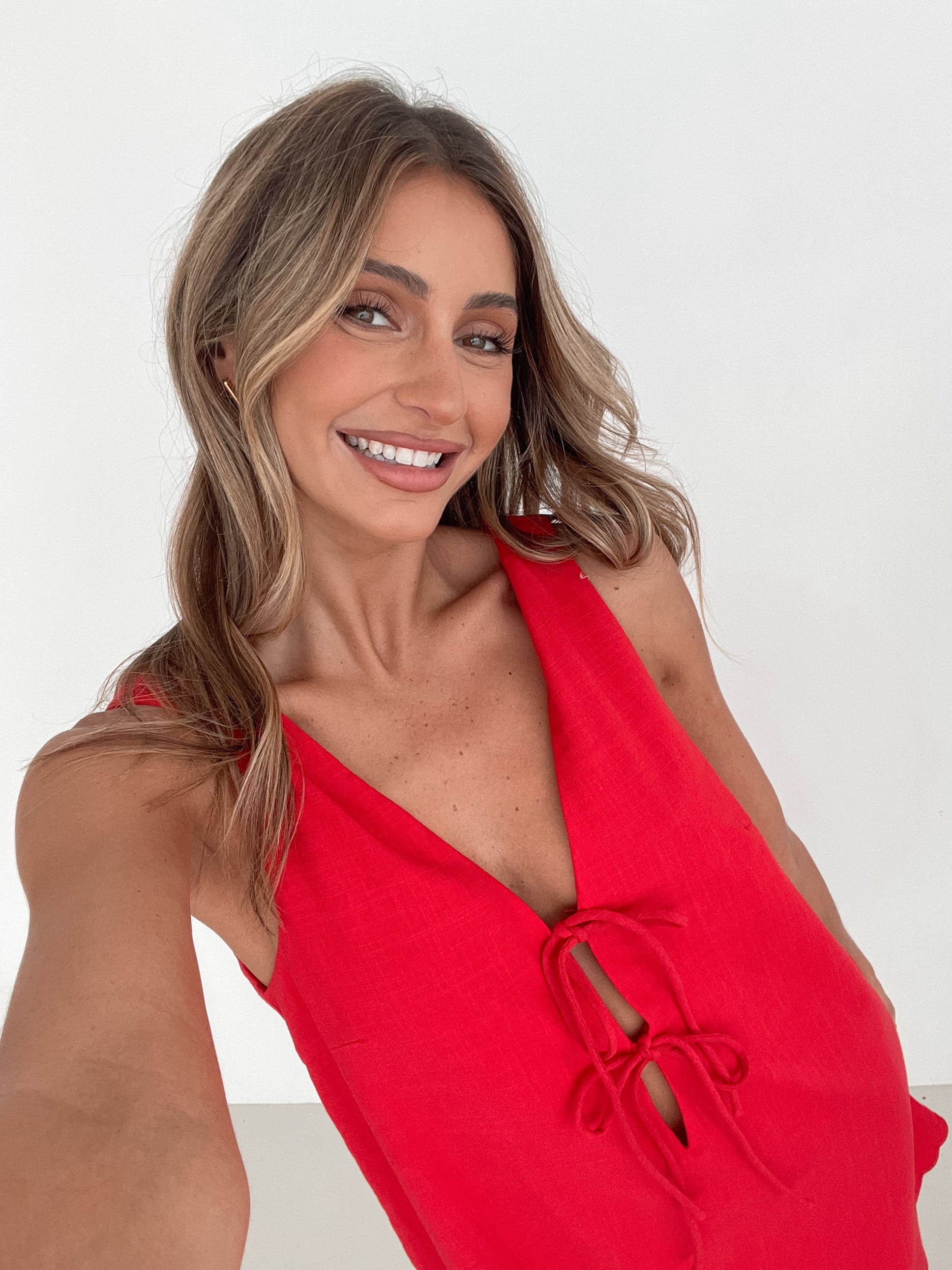 A woman with long, wavy blonde hair stands indoors in front of a plain white wall, with a green leafy plant beside her. She is wearing a sleeveless red dress with a V-neck and two front tie details, similar in style to the previous dress, featuring a scalloped hem. Her hands are gently clasped together in front of her, and she has a subtle smile on her face. She accessorizes with gold hoop earrings and a delicate ring, and her makeup is natural and polished.