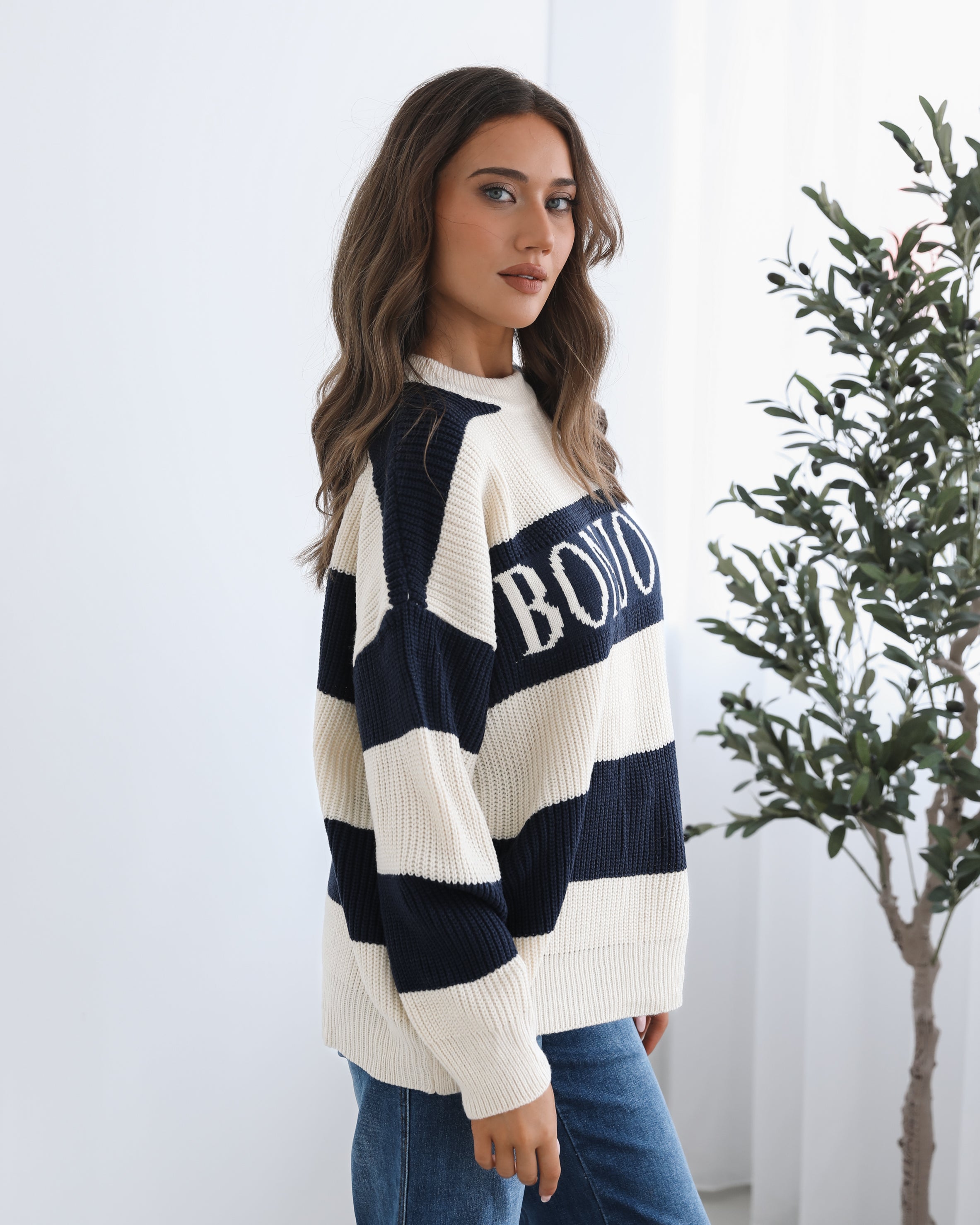 A woman with long, wavy brown hair is seated on a wooden chair with a woven backrest, positioned indoors against a plain white wall and next to a tall green leafy plant. She is wearing a cozy, oversized knitted sweater with wide horizontal stripes in cream and navy blue. The sweater has the word "BONJOUR" written across the chest in large, bold letters. She pairs the sweater with loose-fitting blue jeans. Her pose is relaxed, with her hands resting on her knees, and she gazes softly toward the camera with a