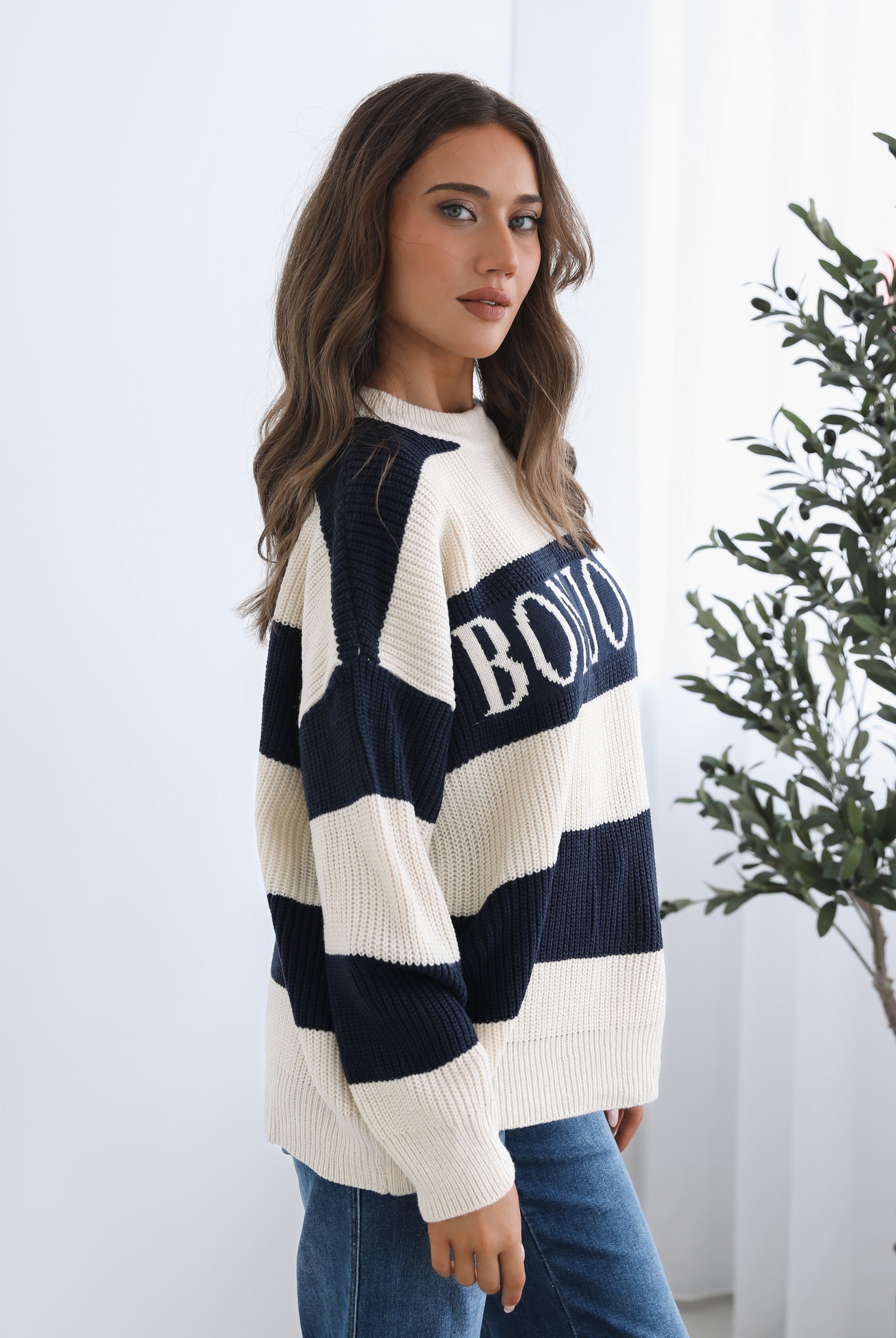 A woman with long, wavy brown hair is seated on a wooden chair with a woven backrest, positioned indoors against a plain white wall and next to a tall green leafy plant. She is wearing a cozy, oversized knitted sweater with wide horizontal stripes in cream and navy blue. The sweater has the word "BONJOUR" written across the chest in large, bold letters. She pairs the sweater with loose-fitting blue jeans. Her pose is relaxed, with her hands resting on her knees, and she gazes softly toward the camera with a