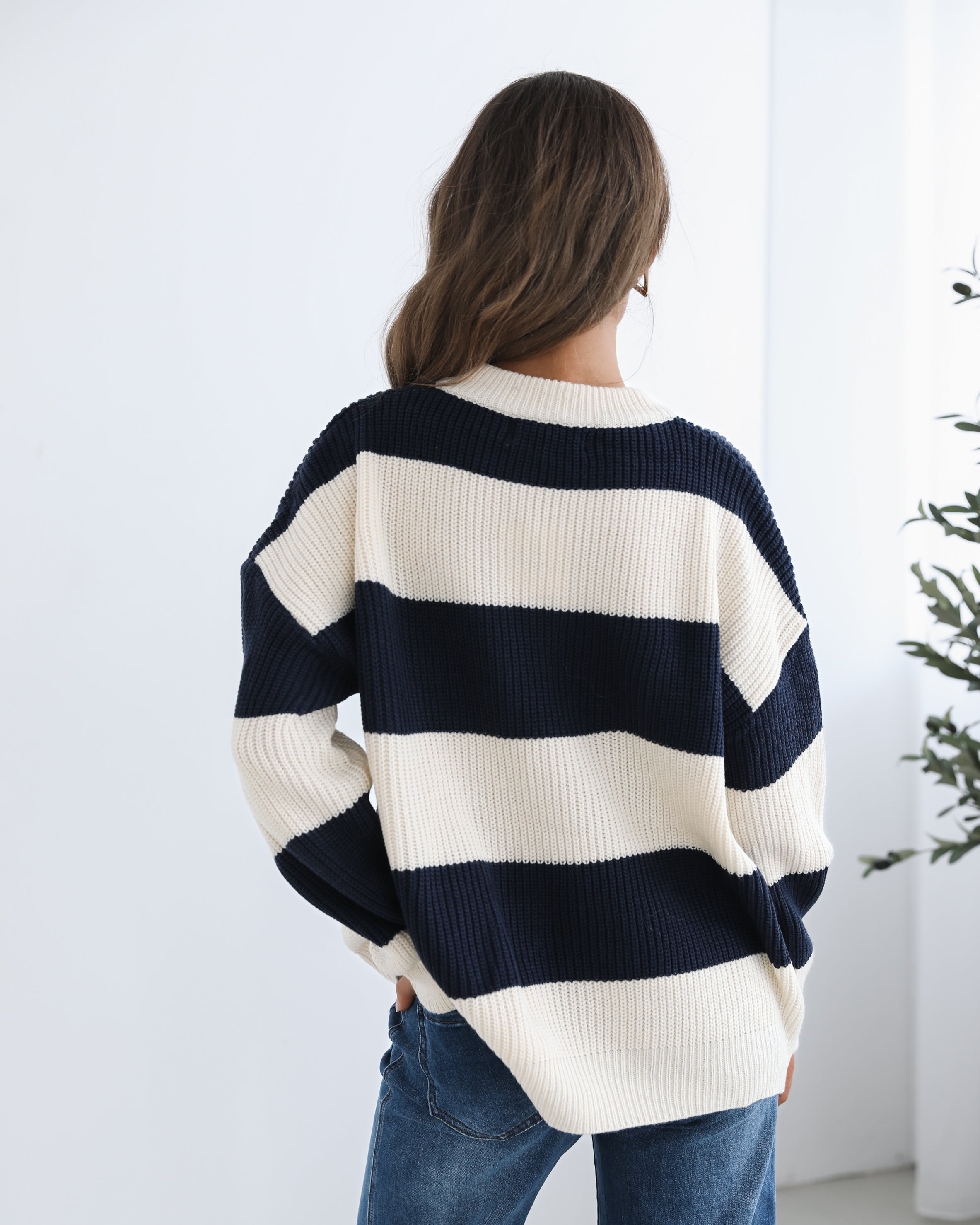 A woman with long, wavy brown hair is seated on a wooden chair with a woven backrest, positioned indoors against a plain white wall and next to a tall green leafy plant. She is wearing a cozy, oversized knitted sweater with wide horizontal stripes in cream and navy blue. The sweater has the word "BONJOUR" written across the chest in large, bold letters. She pairs the sweater with loose-fitting blue jeans. Her pose is relaxed, with her hands resting on her knees, and she gazes softly toward the camera with a