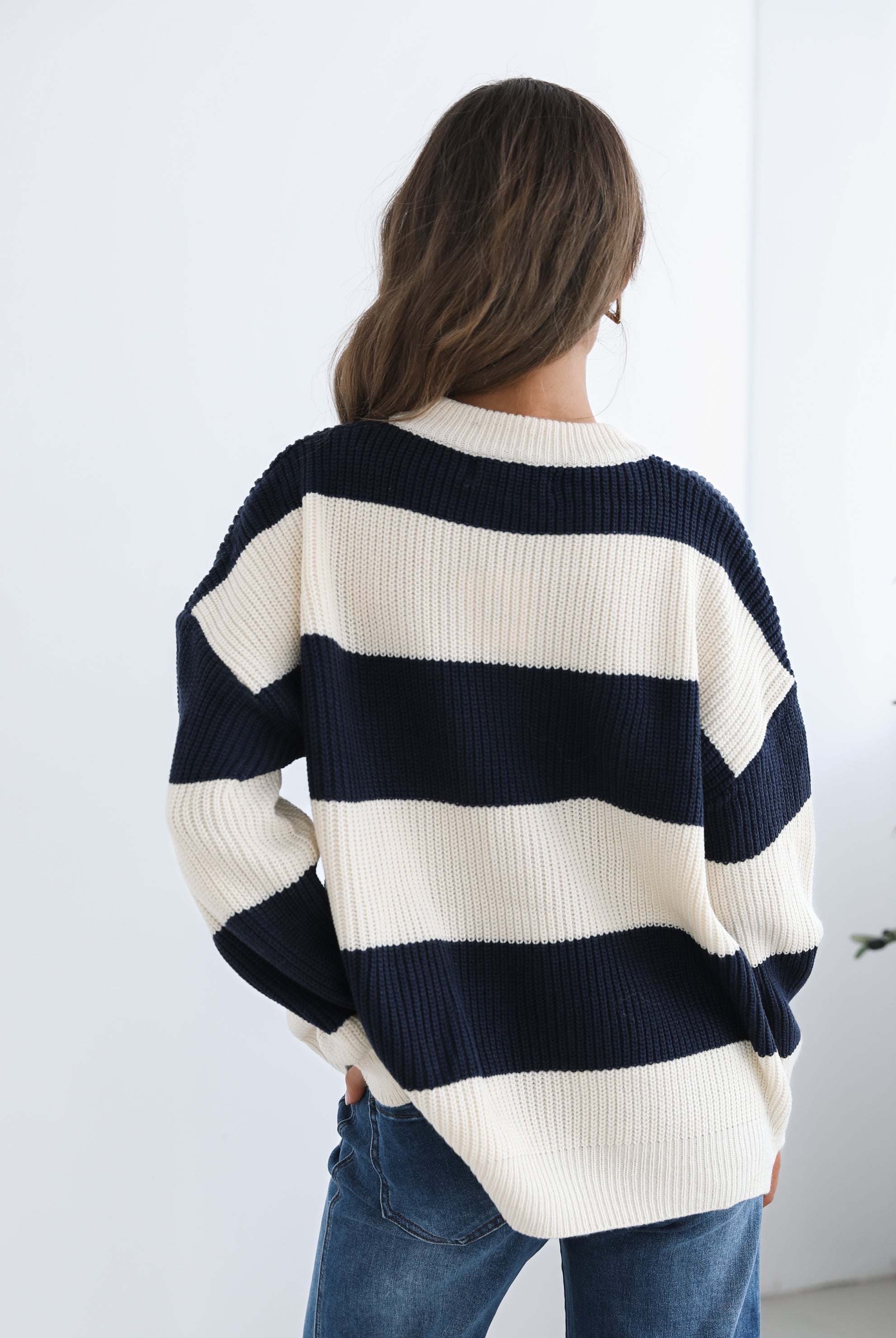 A woman with long, wavy brown hair is seated on a wooden chair with a woven backrest, positioned indoors against a plain white wall and next to a tall green leafy plant. She is wearing a cozy, oversized knitted sweater with wide horizontal stripes in cream and navy blue. The sweater has the word "BONJOUR" written across the chest in large, bold letters. She pairs the sweater with loose-fitting blue jeans. Her pose is relaxed, with her hands resting on her knees, and she gazes softly toward the camera with a