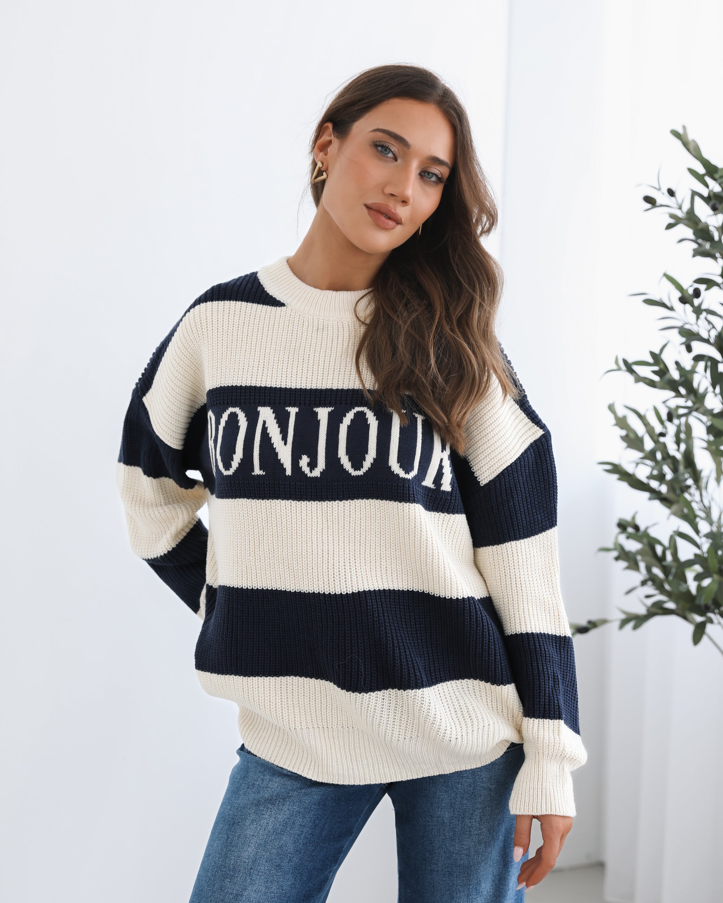 A woman with long, wavy brown hair is seated on a wooden chair with a woven backrest, positioned indoors against a plain white wall and next to a tall green leafy plant. She is wearing a cozy, oversized knitted sweater with wide horizontal stripes in cream and navy blue. The sweater has the word "BONJOUR" written across the chest in large, bold letters. She pairs the sweater with loose-fitting blue jeans. Her pose is relaxed, with her hands resting on her knees, and she gazes softly toward the camera with a
