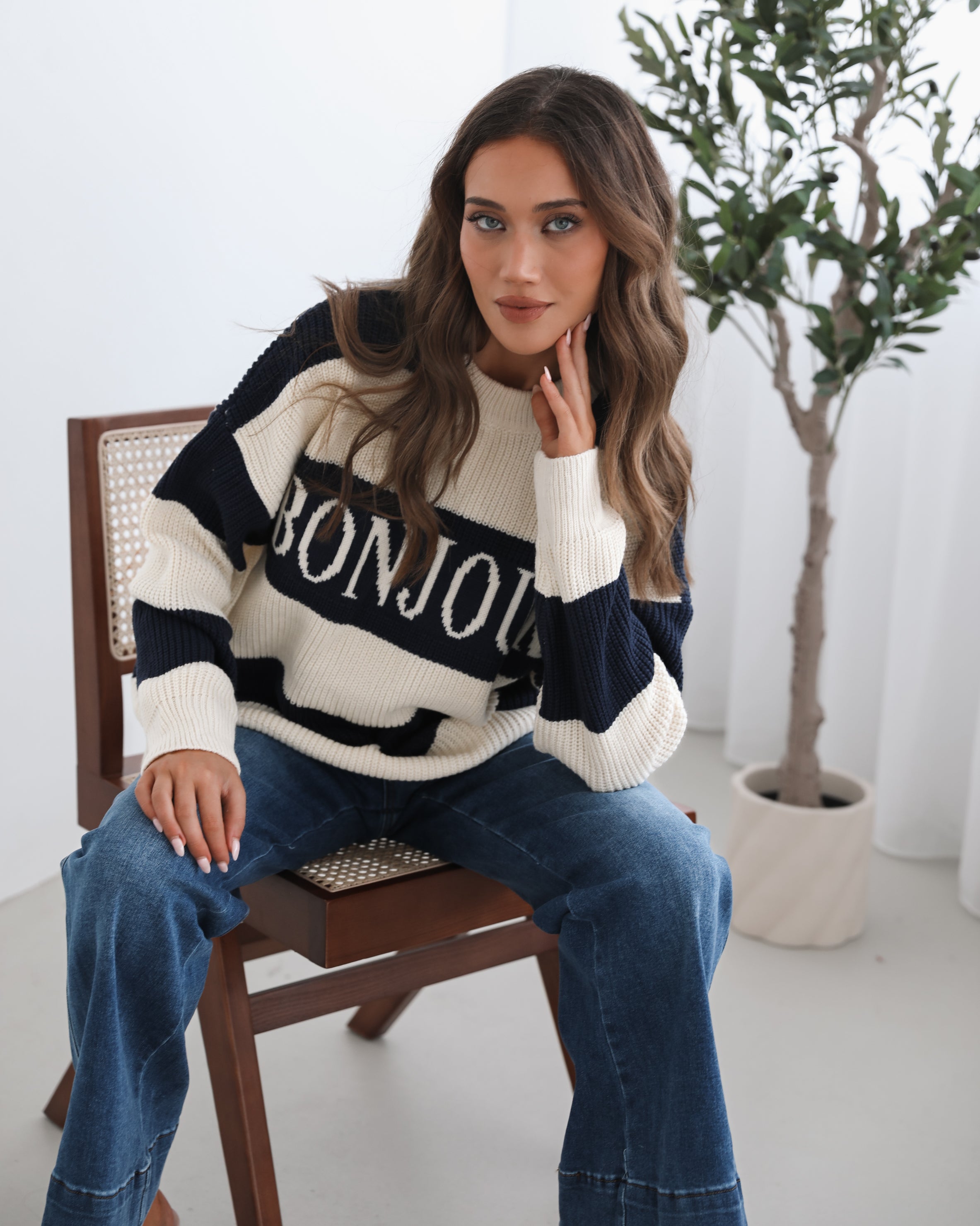 A woman with long, wavy brown hair is seated on a wooden chair with a woven backrest, positioned indoors against a plain white wall and next to a tall green leafy plant. She is wearing a cozy, oversized knitted sweater with wide horizontal stripes in cream and navy blue. The sweater has the word "BONJOUR" written across the chest in large, bold letters. She pairs the sweater with loose-fitting blue jeans. Her pose is relaxed, with her hands resting on her knees, and she gazes softly toward the camera with a