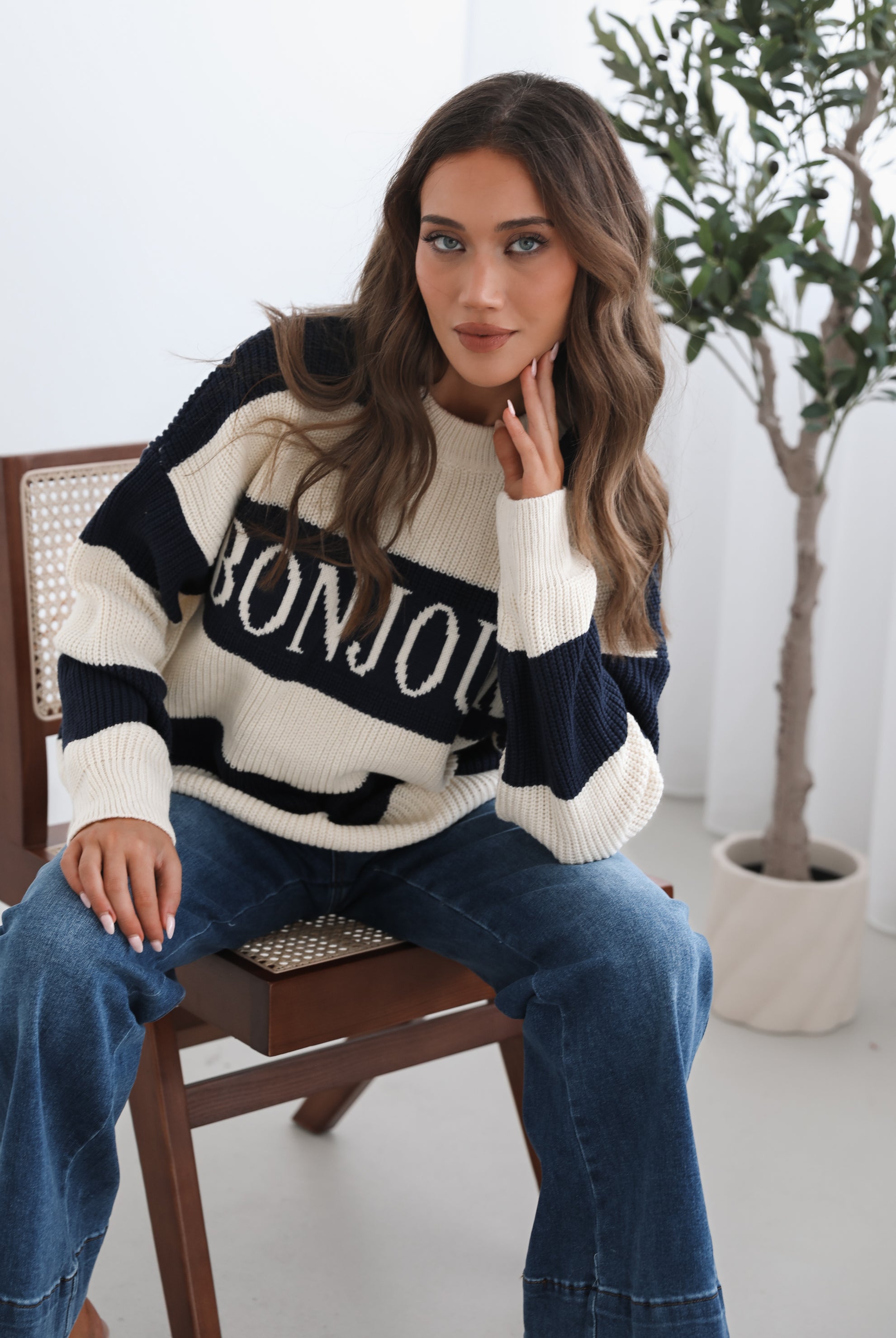 A woman with long, wavy brown hair is seated on a wooden chair with a woven backrest, positioned indoors against a plain white wall and next to a tall green leafy plant. She is wearing a cozy, oversized knitted sweater with wide horizontal stripes in cream and navy blue. The sweater has the word "BONJOUR" written across the chest in large, bold letters. She pairs the sweater with loose-fitting blue jeans. Her pose is relaxed, with her hands resting on her knees, and she gazes softly toward the camera with a