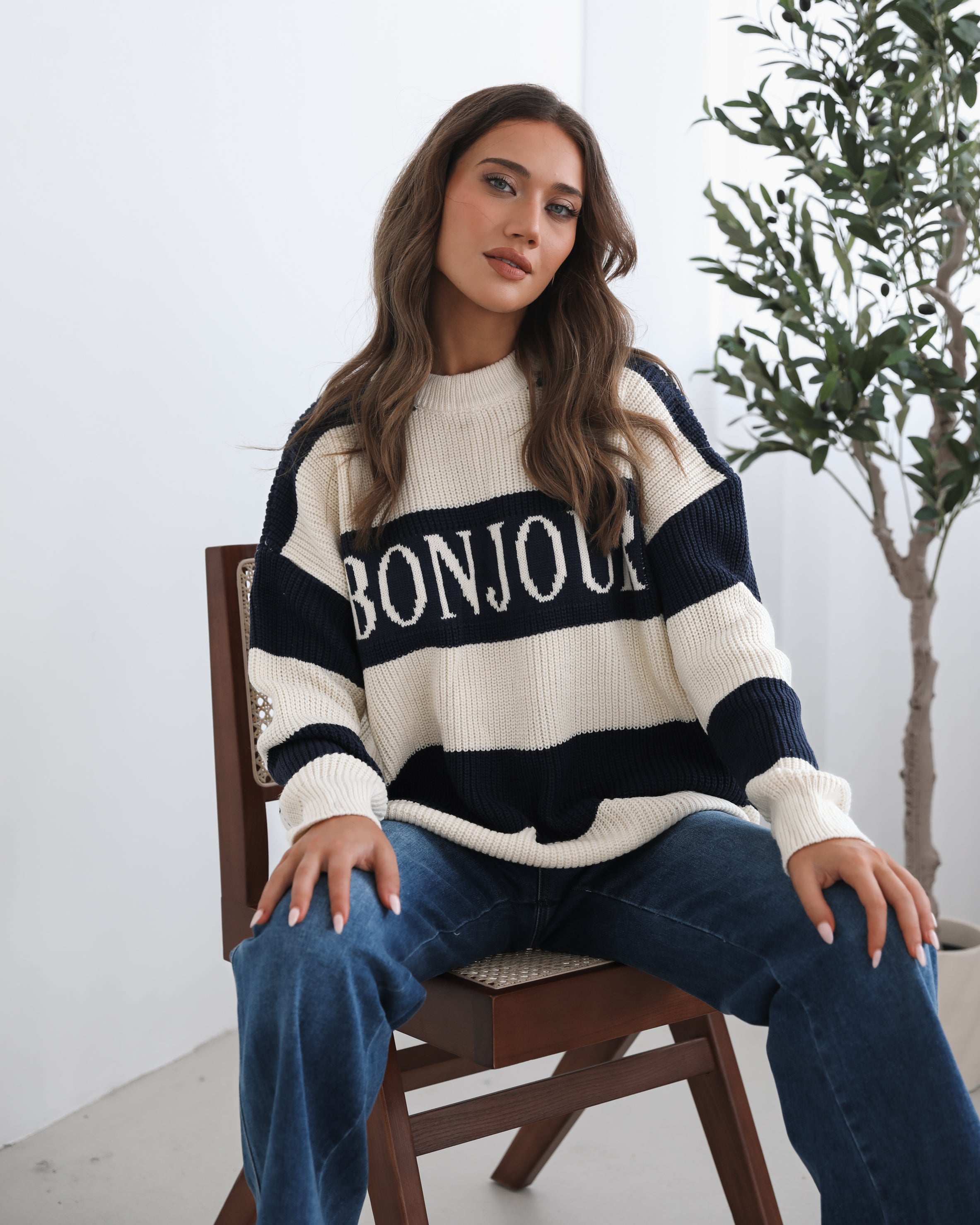 A woman with long, wavy brown hair is seated on a wooden chair with a woven backrest, positioned indoors against a plain white wall and next to a tall green leafy plant. She is wearing a cozy, oversized knitted sweater with wide horizontal stripes in cream and navy blue. The sweater has the word "BONJOUR" written across the chest in large, bold letters. She pairs the sweater with loose-fitting blue jeans. Her pose is relaxed, with her hands resting on her knees, and she gazes softly toward the camera with a