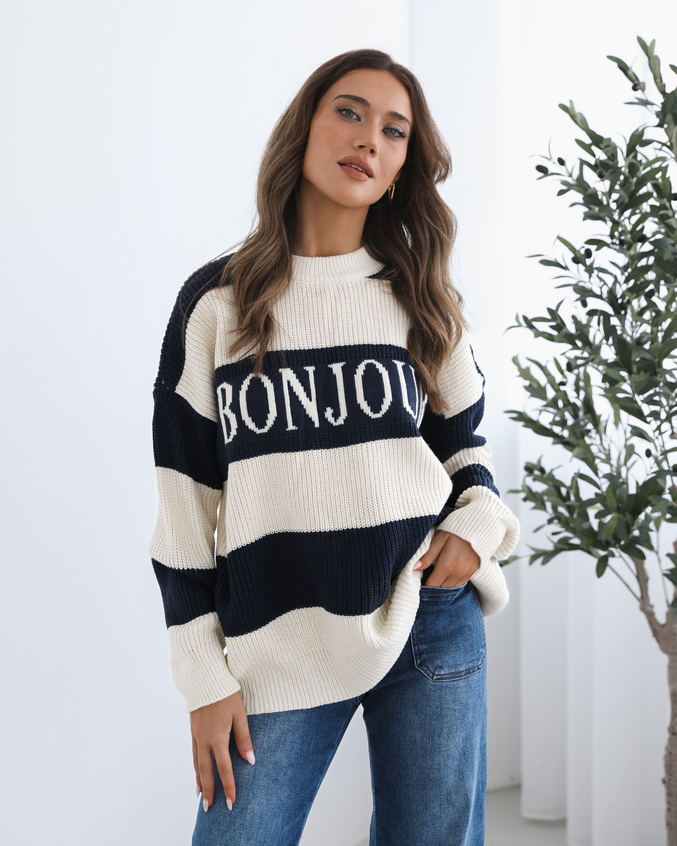 A woman with long, wavy brown hair is seated on a wooden chair with a woven backrest, positioned indoors against a plain white wall and next to a tall green leafy plant. She is wearing a cozy, oversized knitted sweater with wide horizontal stripes in cream and navy blue. The sweater has the word "BONJOUR" written across the chest in large, bold letters. She pairs the sweater with loose-fitting blue jeans. Her pose is relaxed, with her hands resting on her knees, and she gazes softly toward the camera with a