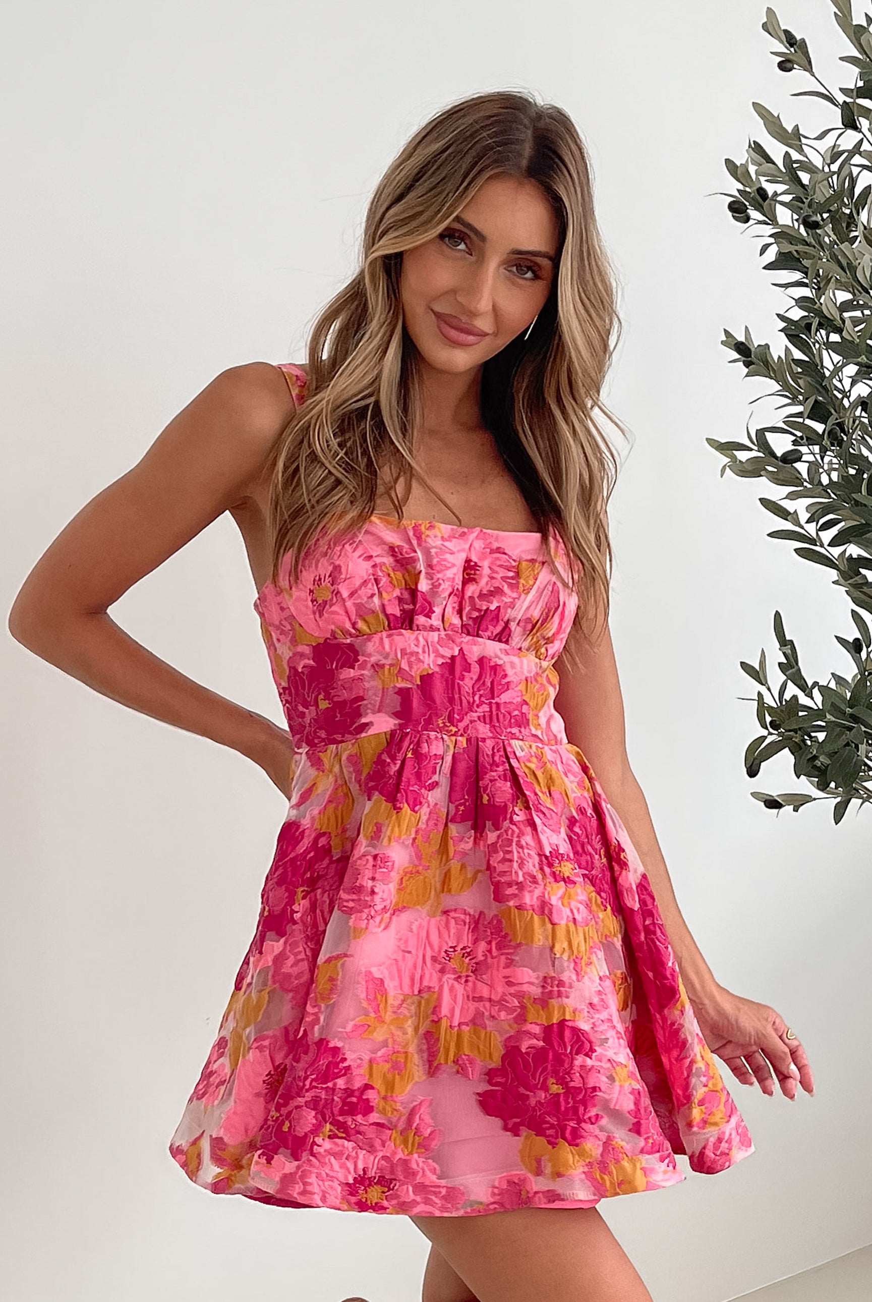 A young woman with long, wavy blonde hair stands indoors against a simple white wall, with a tall leafy plant nearby. She is wearing a sleeveless, strapless summer dress featuring a vibrant floral pattern in shades of pink, magenta, and yellow. The dress has a fitted bodice with a square neckline and a flared skirt that adds a playful and feminine touch. She has a soft smile and her right hand is gently touching her hair, exuding a cheerful and relaxed vibe.