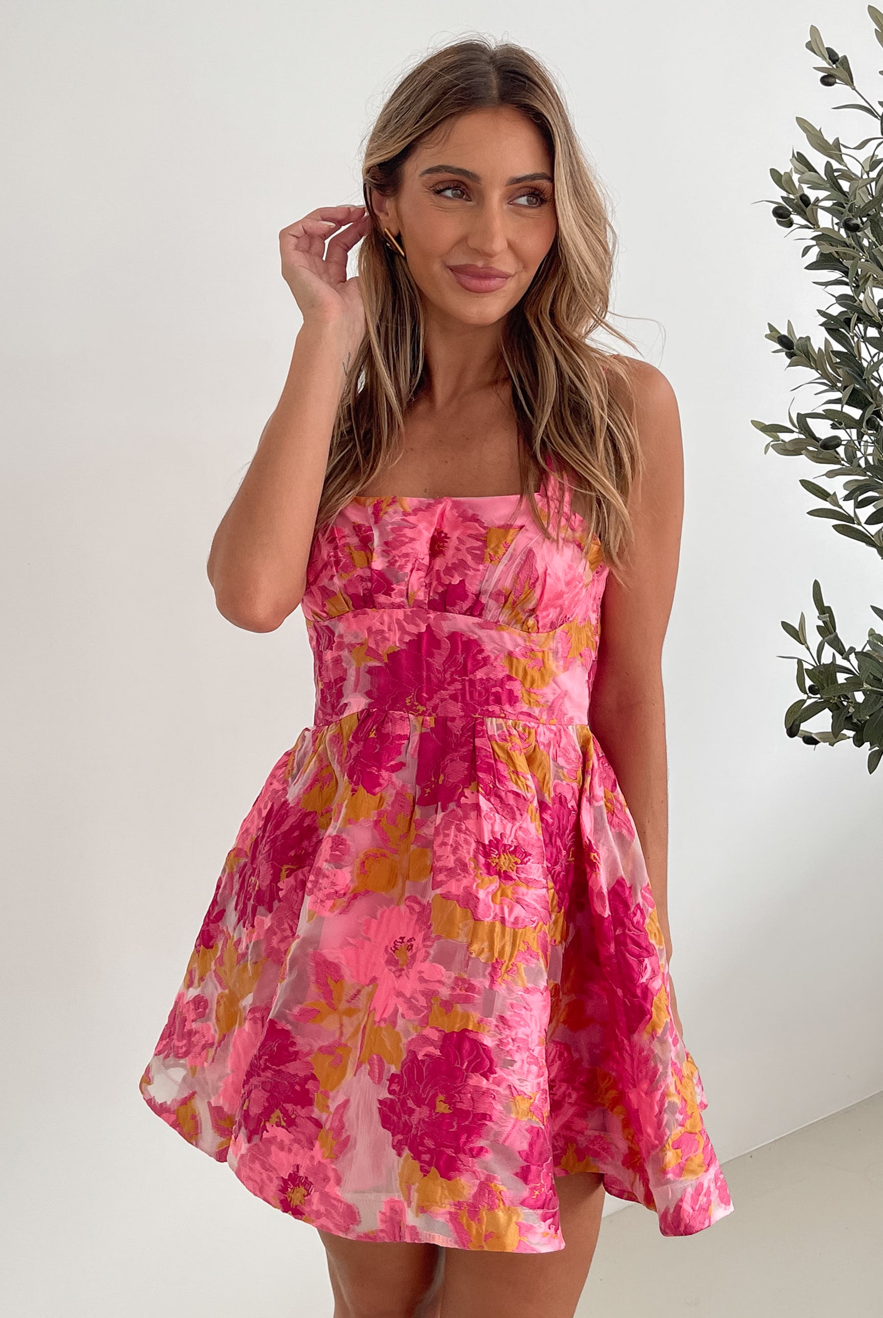 A young woman with long, wavy blonde hair stands indoors against a simple white wall, with a tall leafy plant nearby. She is wearing a sleeveless, strapless summer dress featuring a vibrant floral pattern in shades of pink, magenta, and yellow. The dress has a fitted bodice with a square neckline and a flared skirt that adds a playful and feminine touch. She has a soft smile and her right hand is gently touching her hair, exuding a cheerful and relaxed vibe.