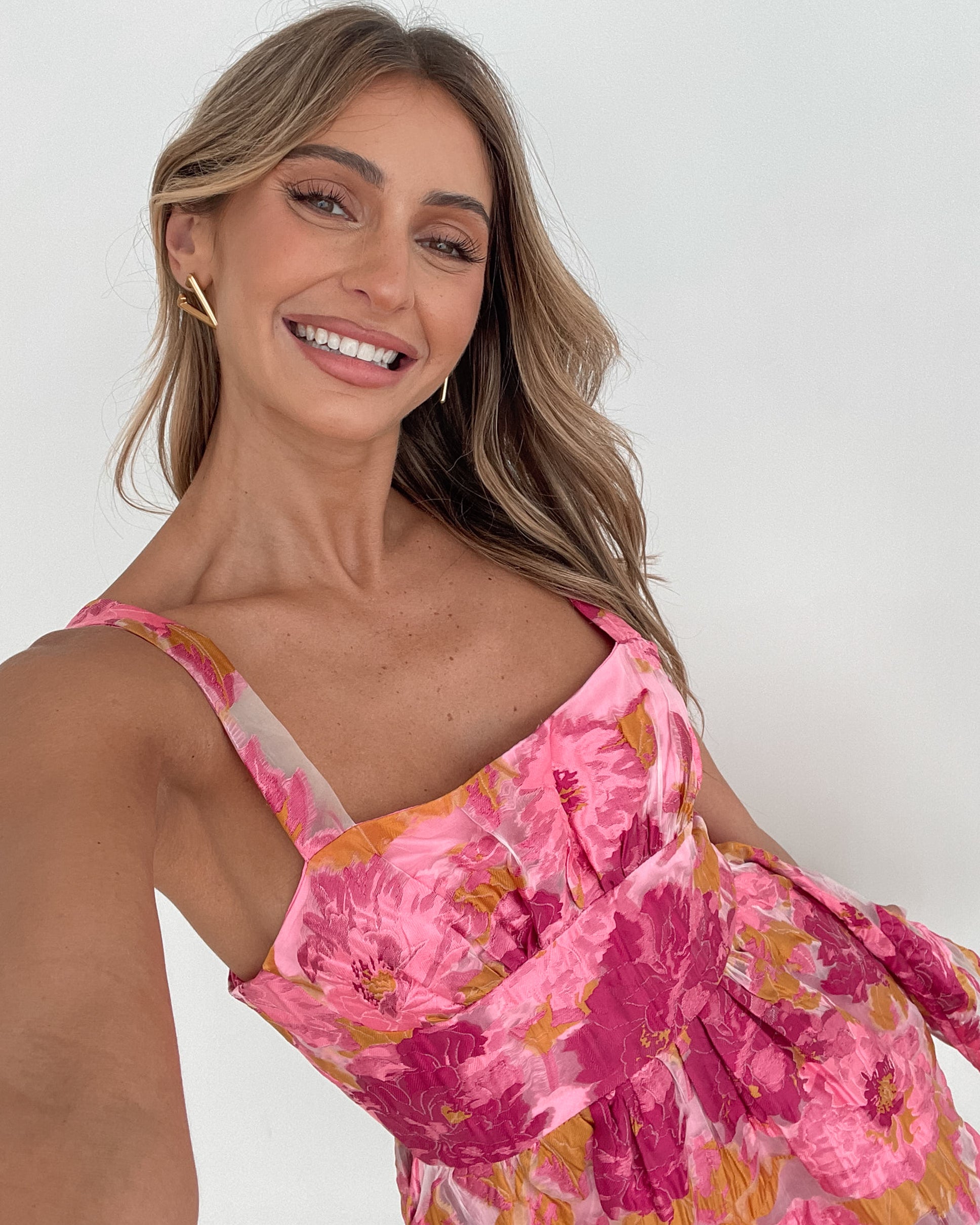 A young woman with long, wavy blonde hair stands indoors against a simple white wall, with a tall leafy plant nearby. She is wearing a sleeveless, strapless summer dress featuring a vibrant floral pattern in shades of pink, magenta, and yellow. The dress has a fitted bodice with a square neckline and a flared skirt that adds a playful and feminine touch. She has a soft smile and her right hand is gently touching her hair, exuding a cheerful and relaxed vibe.