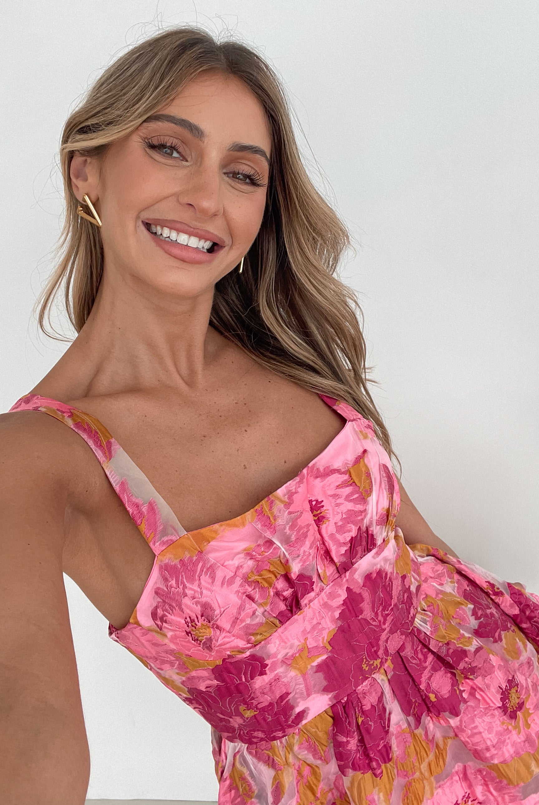 A young woman with long, wavy blonde hair stands indoors against a simple white wall, with a tall leafy plant nearby. She is wearing a sleeveless, strapless summer dress featuring a vibrant floral pattern in shades of pink, magenta, and yellow. The dress has a fitted bodice with a square neckline and a flared skirt that adds a playful and feminine touch. She has a soft smile and her right hand is gently touching her hair, exuding a cheerful and relaxed vibe.