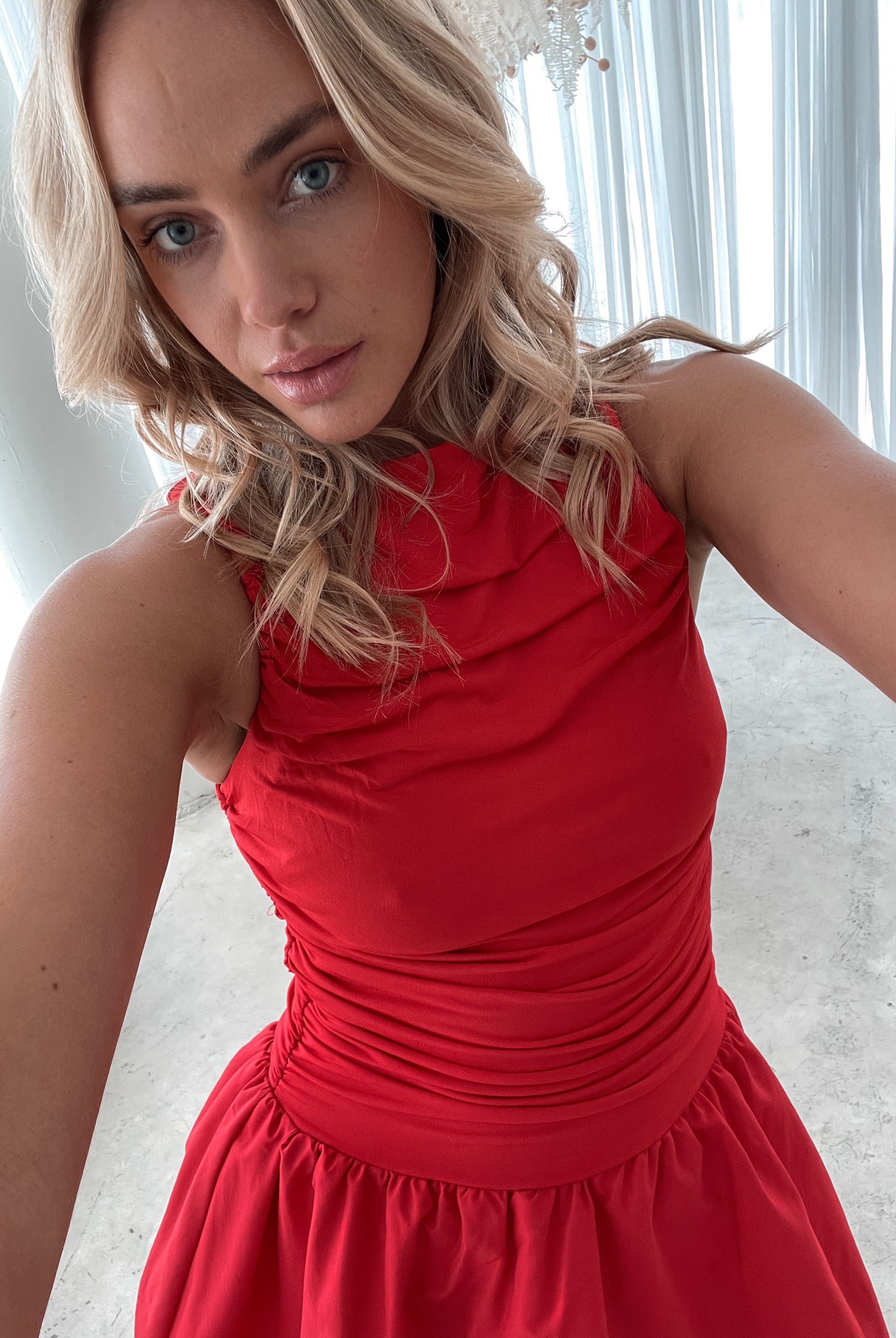 A young woman with blonde hair styled in loose waves stands indoors in front of sheer white curtains. She is wearing a vibrant red sleeveless dress with a fitted, ruched bodice and a playful, bubble-hem skirt. The dress has a high neckline and accentuates her waist, creating a flattering silhouette. She holds the skirt slightly out with one hand, smiling softly with a confident and relaxed expression. The setting and her pose give off an elegant yet fun and youthful vibe.