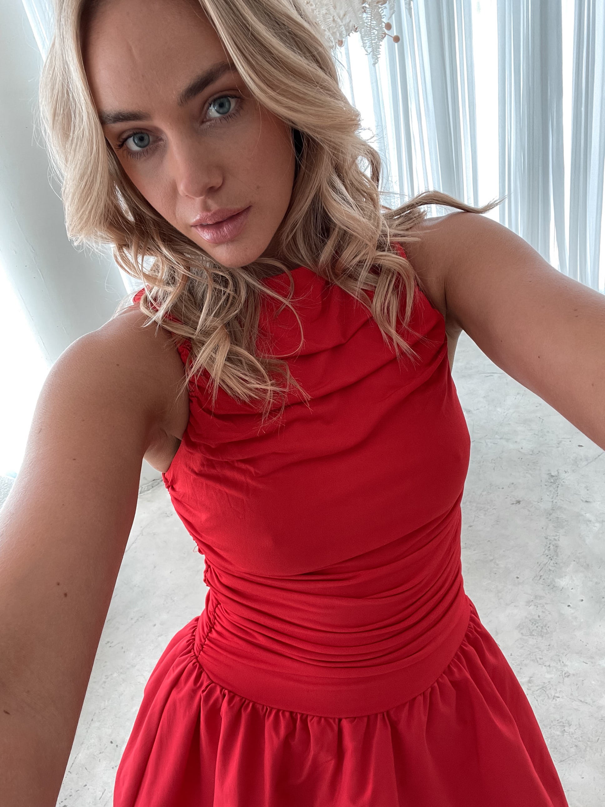 A young woman with blonde hair styled in loose waves stands indoors in front of sheer white curtains. She is wearing a vibrant red sleeveless dress with a fitted, ruched bodice and a playful, bubble-hem skirt. The dress has a high neckline and accentuates her waist, creating a flattering silhouette. She holds the skirt slightly out with one hand, smiling softly with a confident and relaxed expression. The setting and her pose give off an elegant yet fun and youthful vibe.