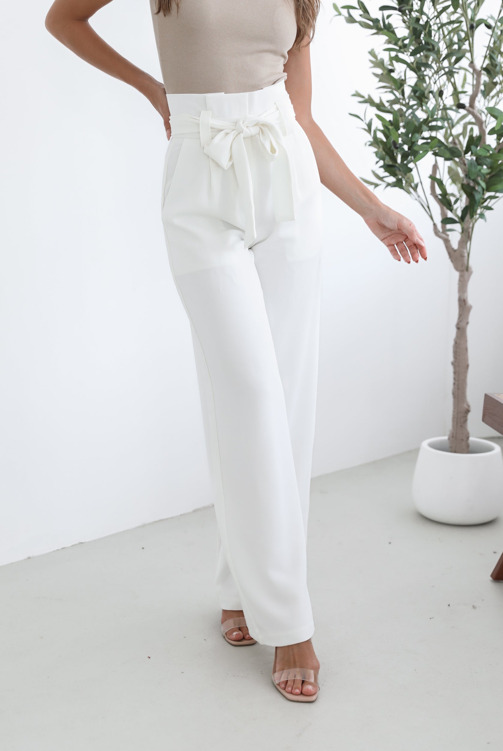 A woman wearing high-waisted, wide-leg white trousers with a paper bag waist and a fabric tie belt, paired with a sleeveless beige ribbed top and nude heels, standing in a minimalistic room with a potted tree and a wooden bench.