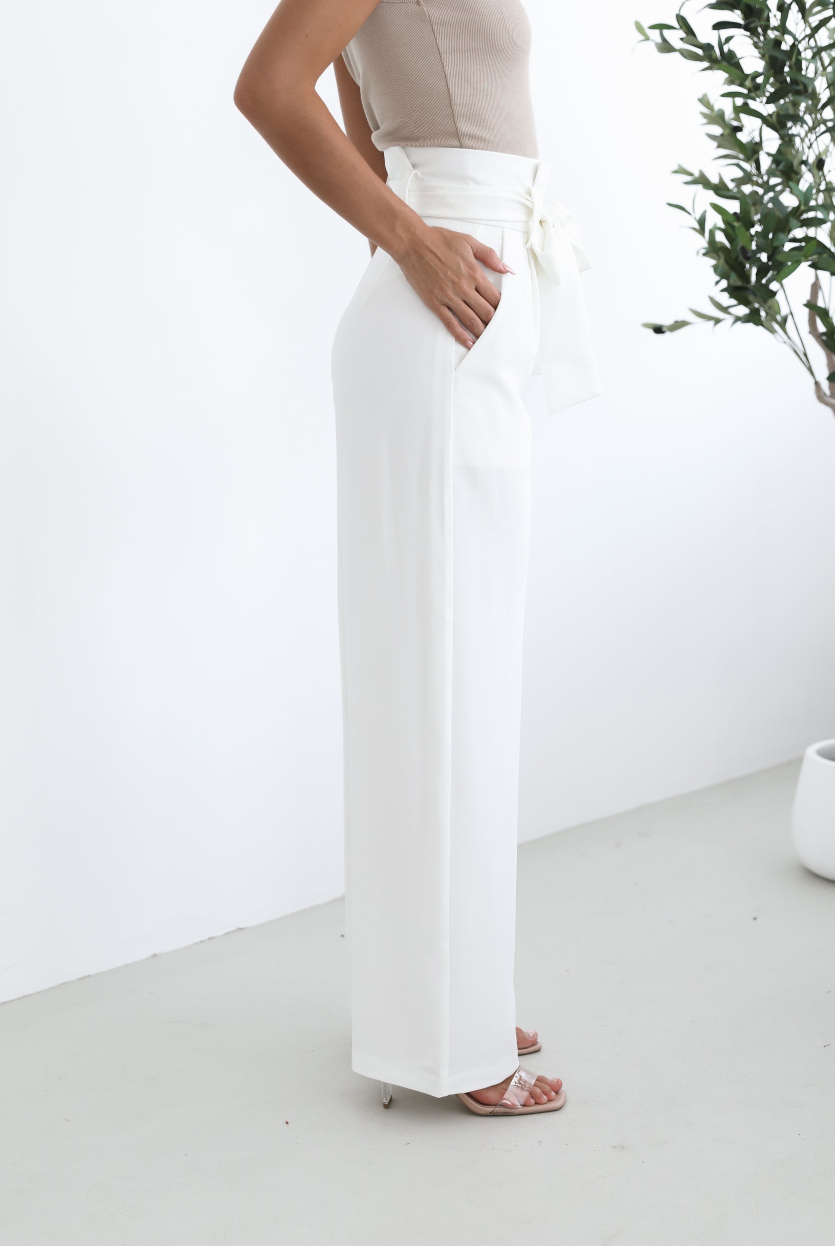 A woman wearing high-waisted, wide-leg white trousers with a paper bag waist and a fabric tie belt, paired with a sleeveless beige ribbed top and nude heels, standing in a minimalistic room with a potted tree and a wooden bench.