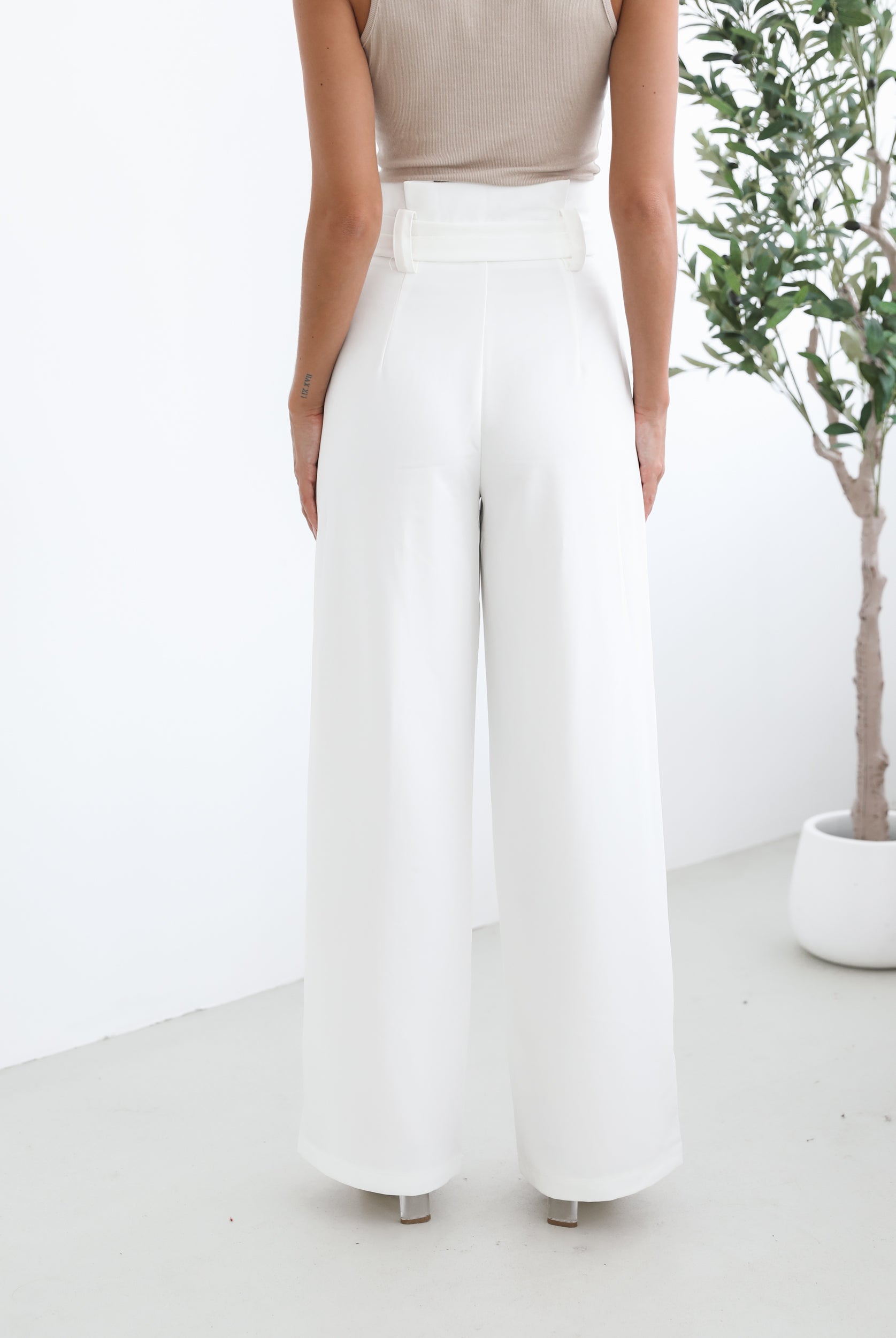 A woman wearing high-waisted, wide-leg white trousers with a paper bag waist and a fabric tie belt, paired with a sleeveless beige ribbed top and nude heels, standing in a minimalistic room with a potted tree and a wooden bench.