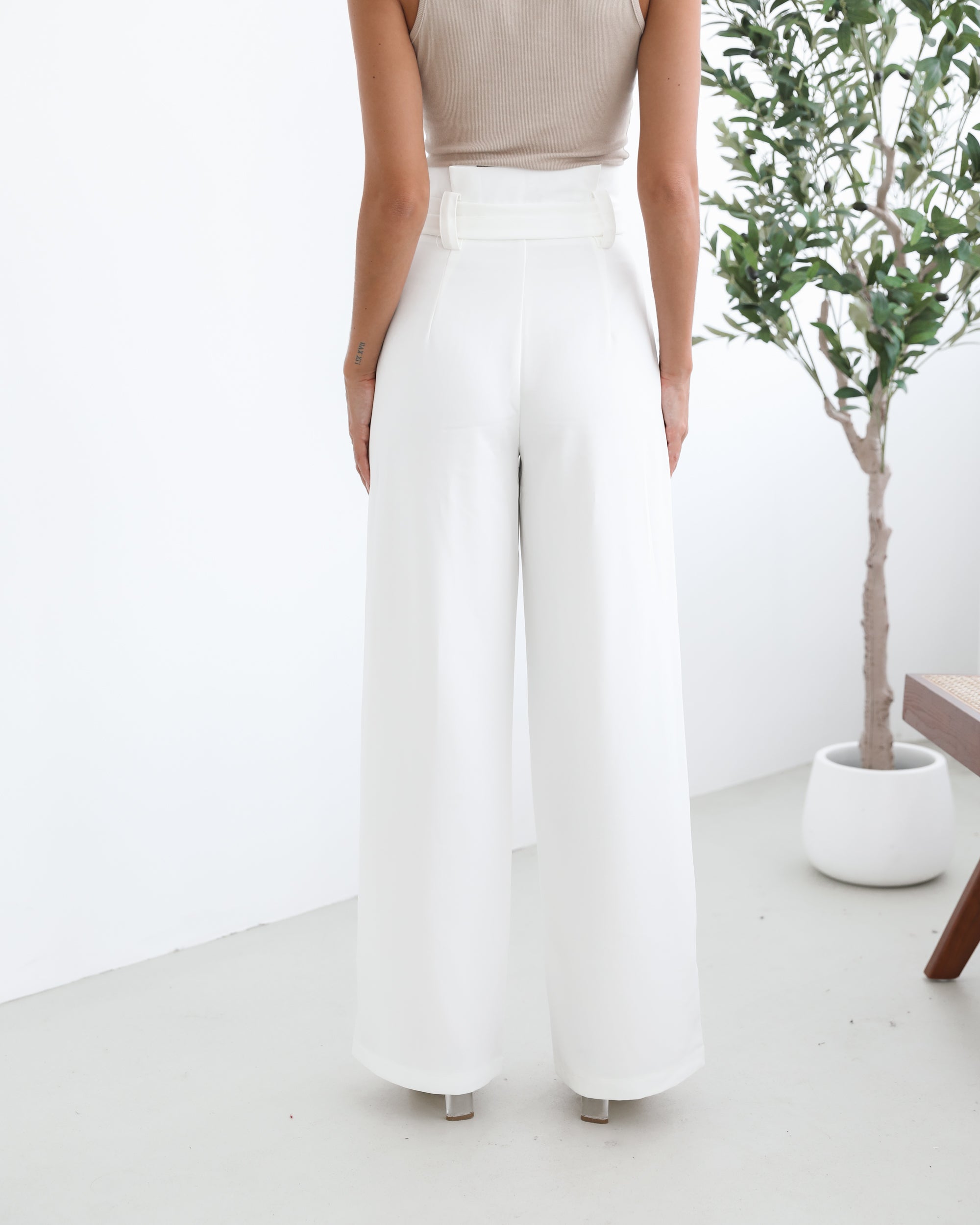 A woman wearing high-waisted, wide-leg white trousers with a paper bag waist and a fabric tie belt, paired with a sleeveless beige ribbed top and nude heels, standing in a minimalistic room with a potted tree and a wooden bench.