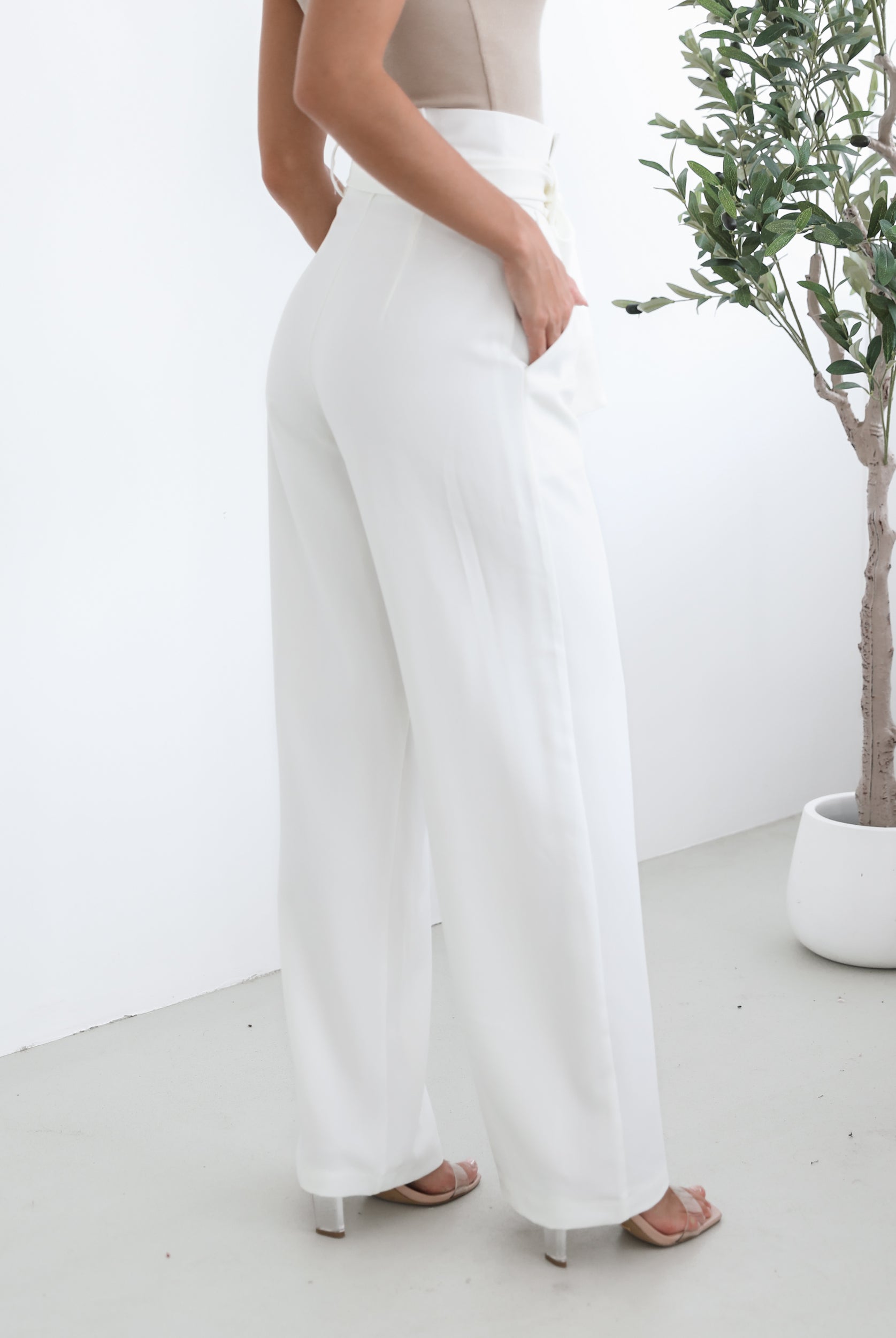 A woman wearing high-waisted, wide-leg white trousers with a paper bag waist and a fabric tie belt, paired with a sleeveless beige ribbed top and nude heels, standing in a minimalistic room with a potted tree and a wooden bench.