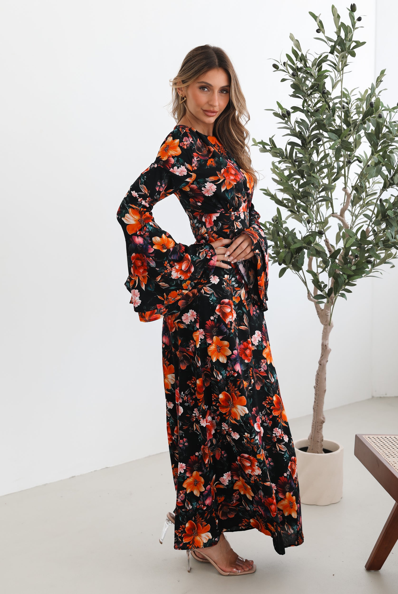 A woman wearing a long, black floral maxi dress with vibrant orange, red, and pink flowers. The dress features long bell sleeves and a fitted waist. She is leaning against a white wall in a minimalistic room with a potted plant nearby.