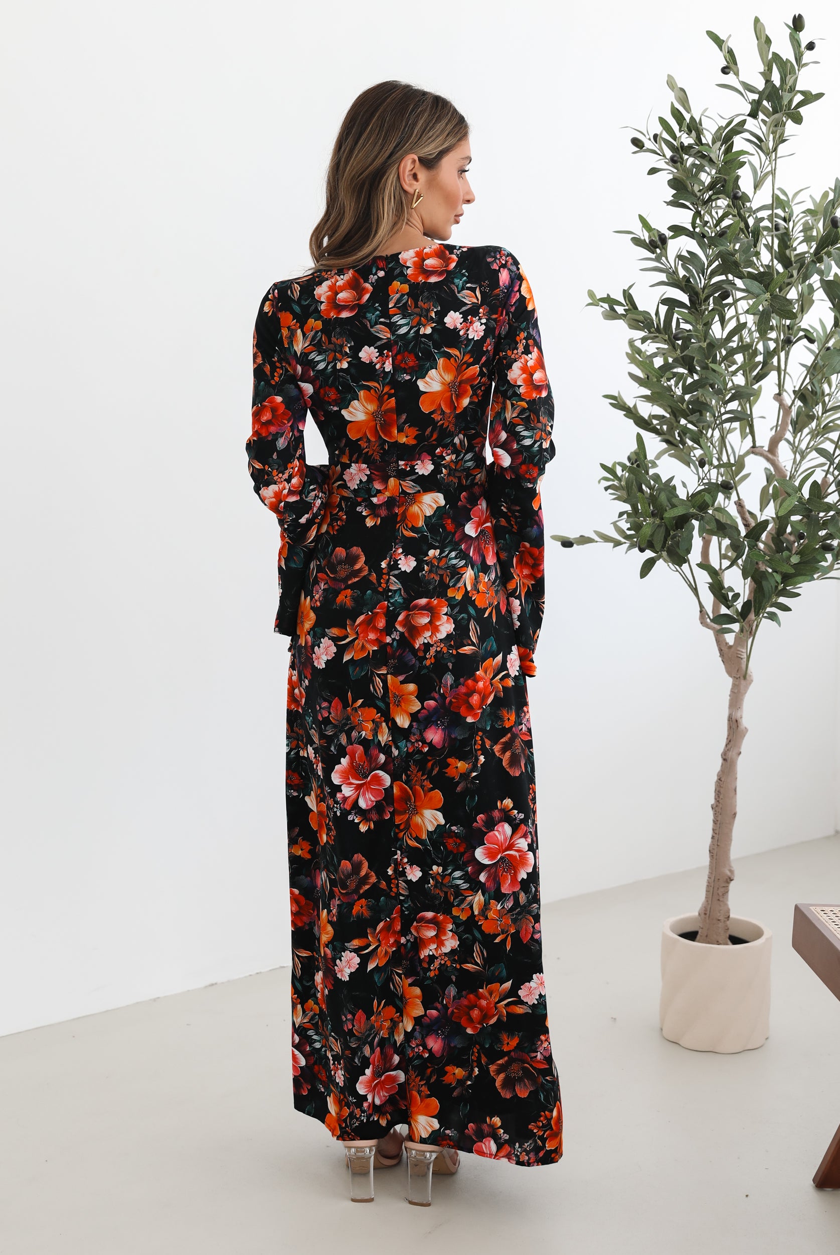A woman wearing a long, black floral maxi dress with vibrant orange, red, and pink flowers. The dress features long bell sleeves and a fitted waist. She is leaning against a white wall in a minimalistic room with a potted plant nearby.