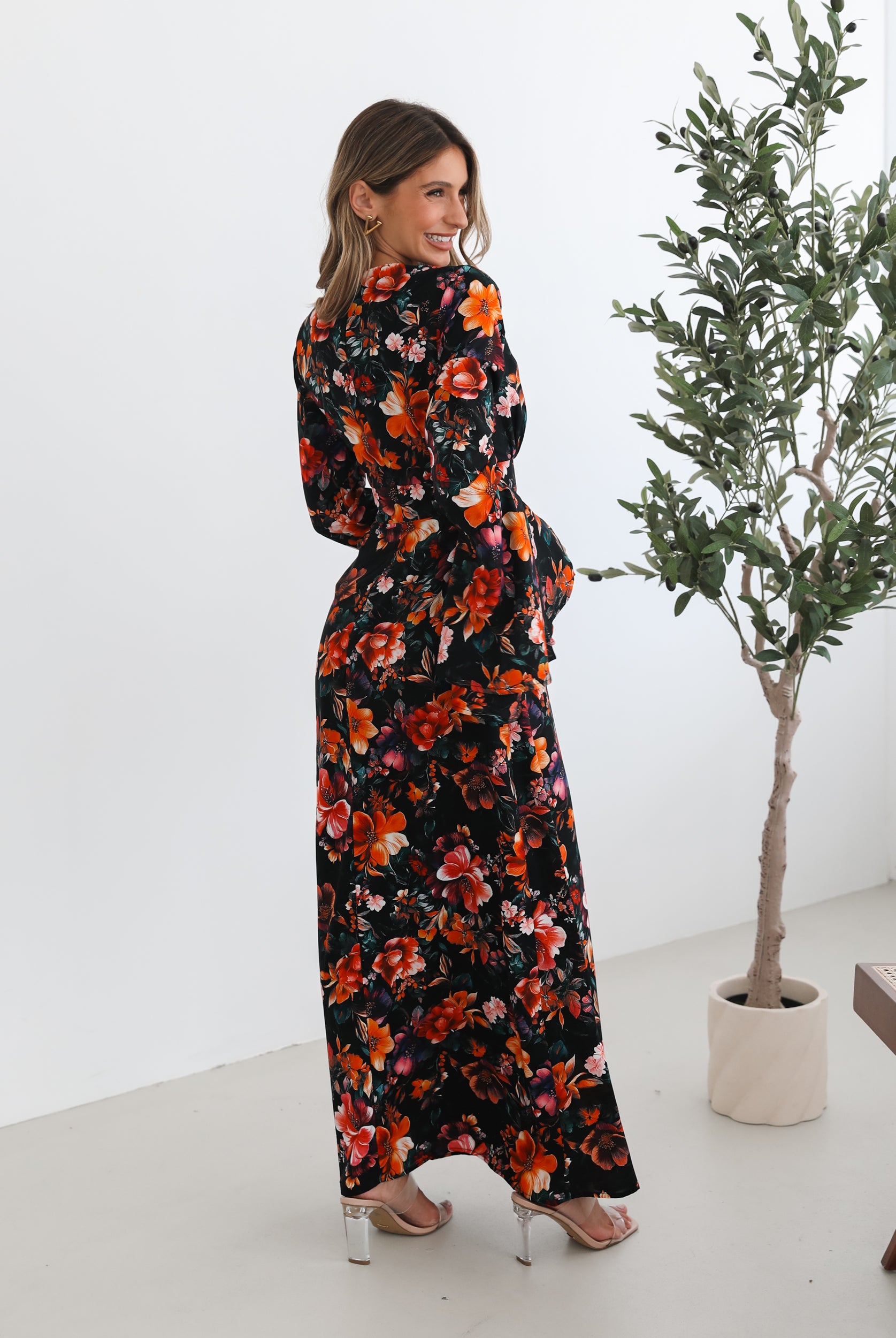 A woman wearing a long, black floral maxi dress with vibrant orange, red, and pink flowers. The dress features long bell sleeves and a fitted waist. She is leaning against a white wall in a minimalistic room with a potted plant nearby.