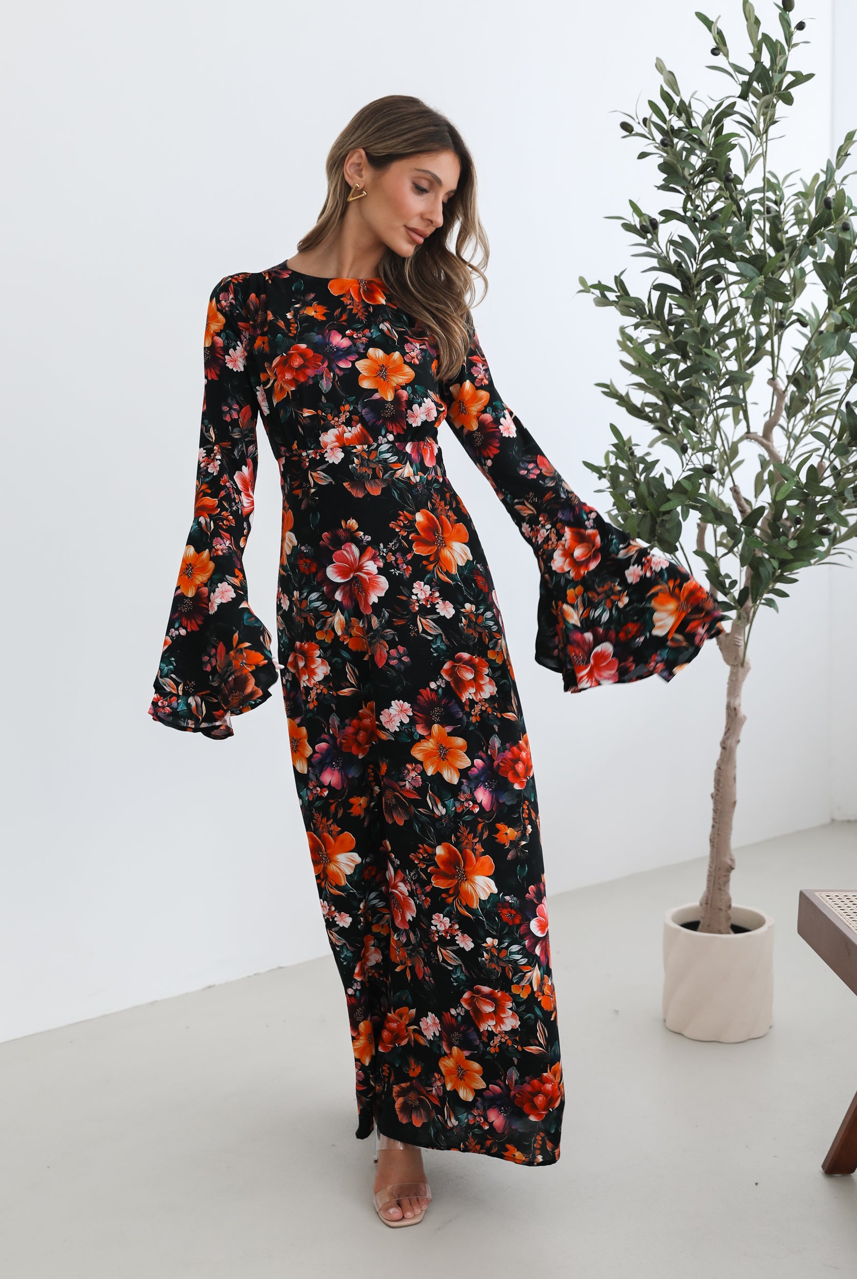 A woman wearing a long, black floral maxi dress with vibrant orange, red, and pink flowers. The dress features long bell sleeves and a fitted waist. She is leaning against a white wall in a minimalistic room with a potted plant nearby.