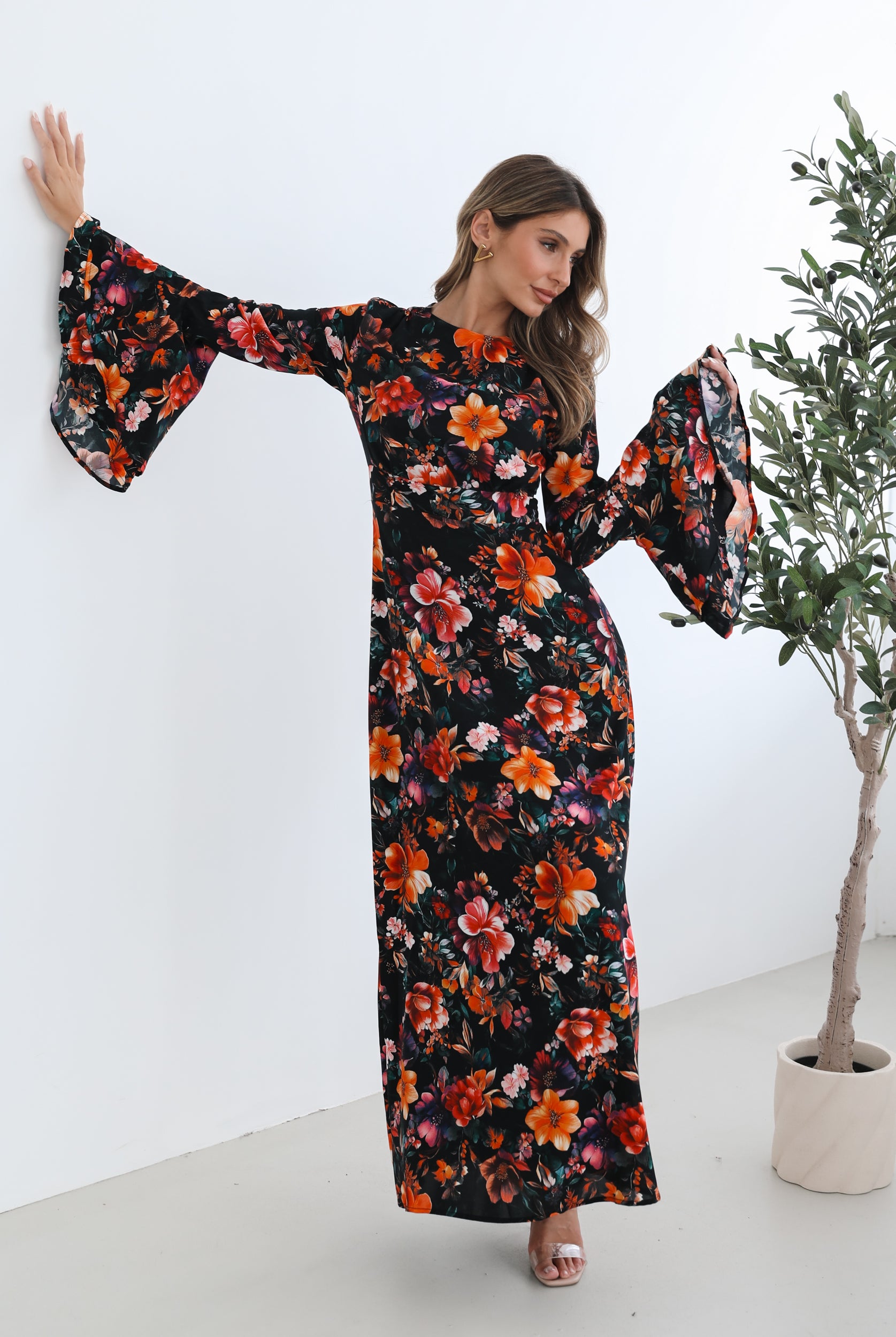 A woman wearing a long, black floral maxi dress with vibrant orange, red, and pink flowers. The dress features long bell sleeves and a fitted waist. She is leaning against a white wall in a minimalistic room with a potted plant nearby.