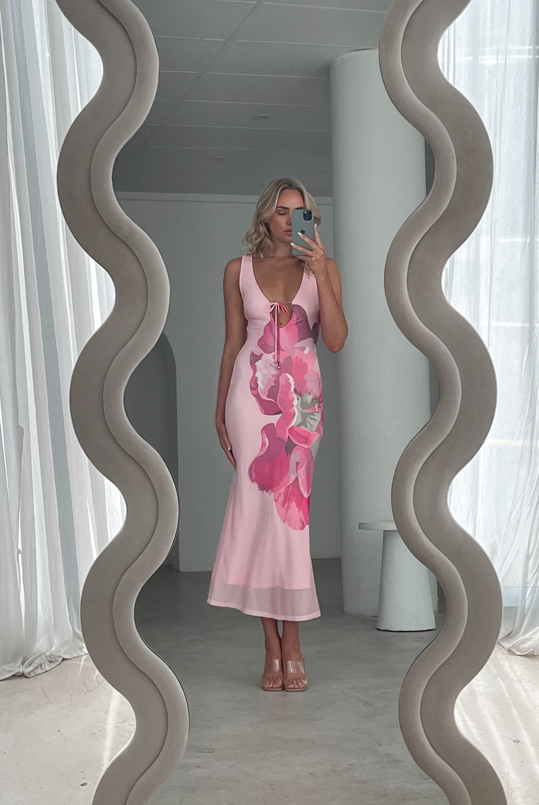 A woman wearing a sleeveless, light pink maxi dress with a deep neckline, front tie detail, and large pink floral print on the front. She stands in a sunlit room with sheer white curtains, beside a modern cream boucle armchair and a small white shelf with minimal decor.