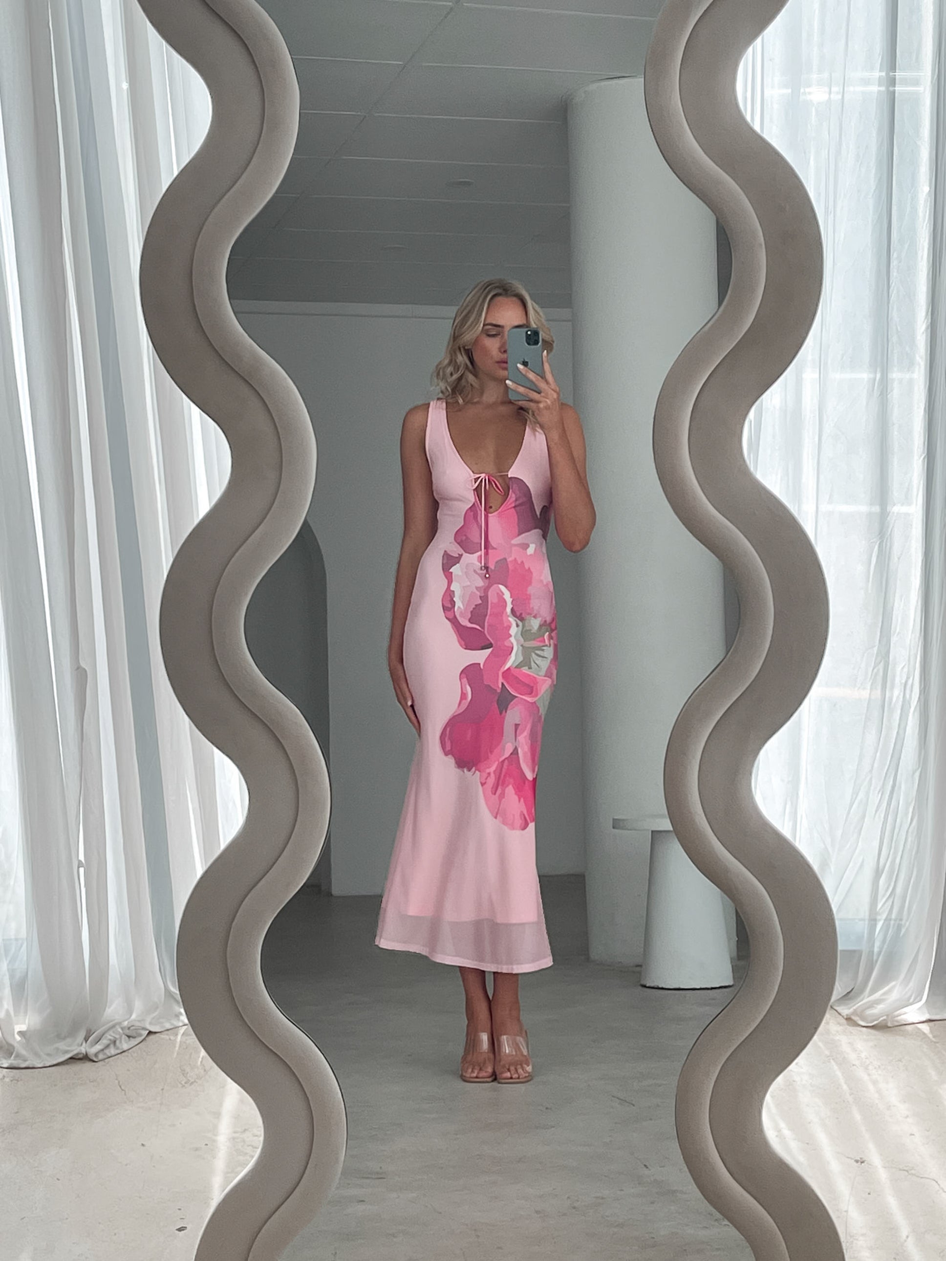 A woman wearing a sleeveless, light pink maxi dress with a deep neckline, front tie detail, and large pink floral print on the front. She stands in a sunlit room with sheer white curtains, beside a modern cream boucle armchair and a small white shelf with minimal decor.