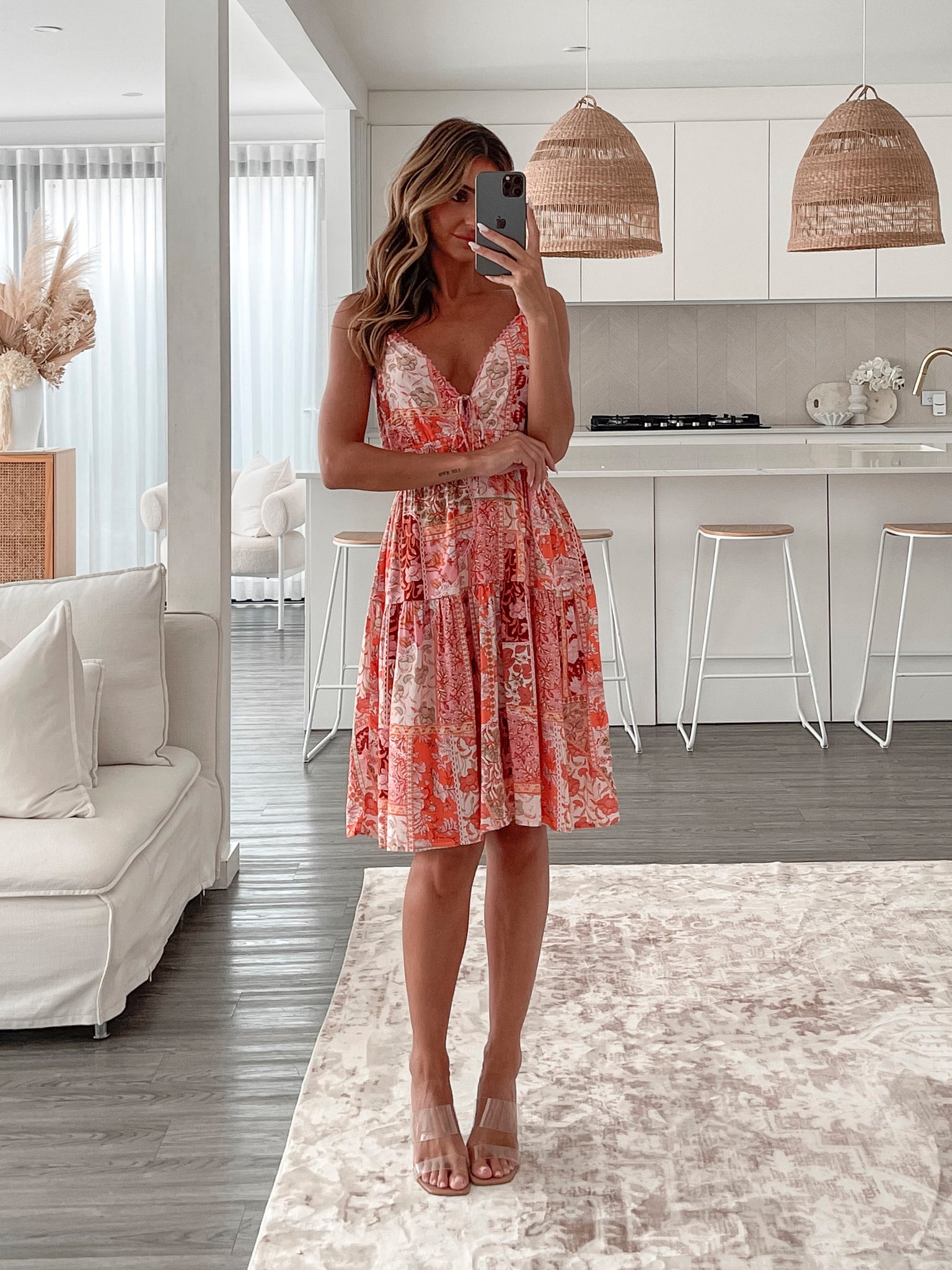 A woman wearing a sleeveless, knee-length summer dress with a deep V-neckline and a flowy silhouette, featuring a vibrant patchwork floral print in shades of orange, pink, and red. She stands indoors near sheer white curtains and a white cabinet with decorative items.