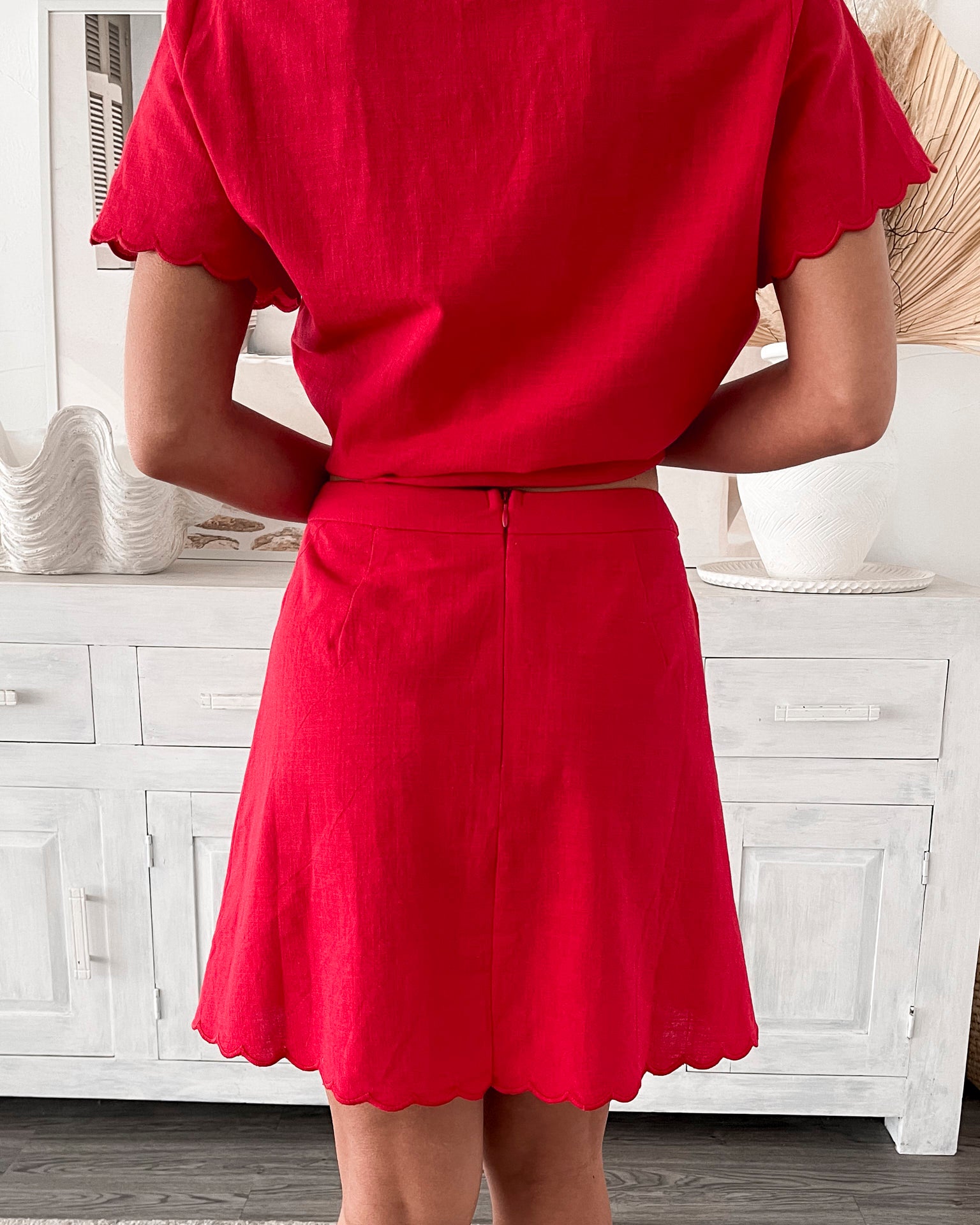 A woman wearing a short, bright red dress with short flutter sleeves and a lace-up front detail. She stands in a stylish room with a white wooden sideboard, neutral decor including dried pampas grass, a woven basket, and a large shell-shaped bowl.