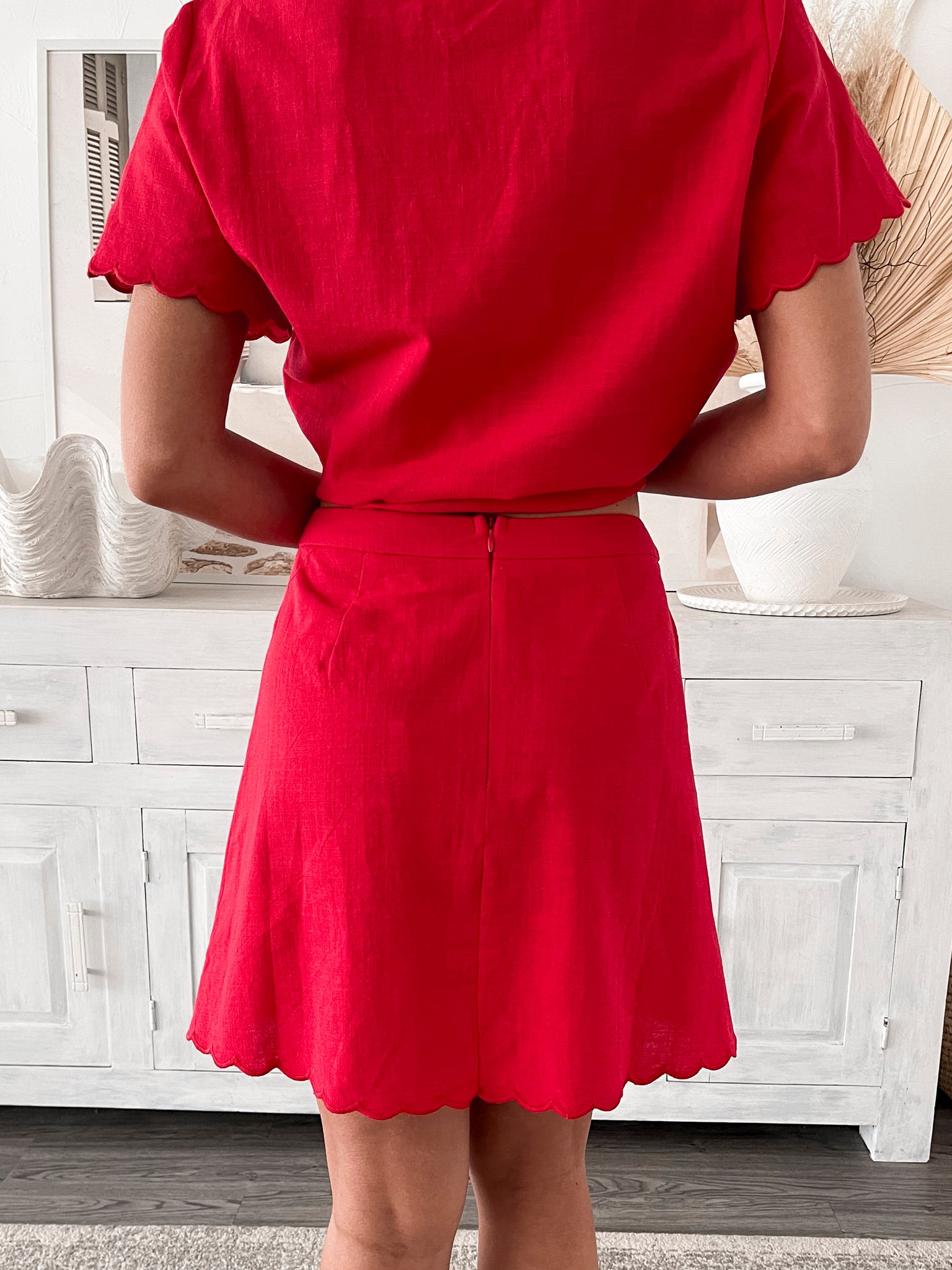 A woman wearing a short, bright red dress with short flutter sleeves and a lace-up front detail. She stands in a stylish room with a white wooden sideboard, neutral decor including dried pampas grass, a woven basket, and a large shell-shaped bowl.
