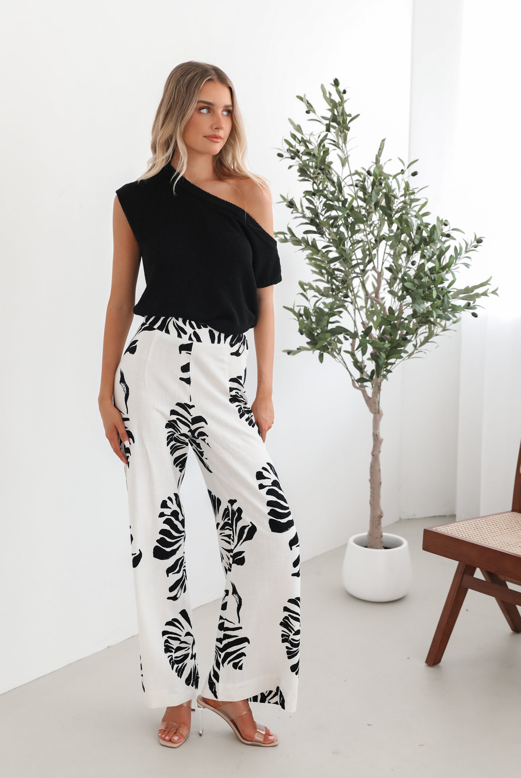 A woman sitting on a wooden chair wearing a black off-the-shoulder top paired with high-waisted white wide-leg pants featuring bold black tropical leaf prints. She completes the look with nude heels and is posed in a minimalistic room with white curtains and a potted plant in the background.
