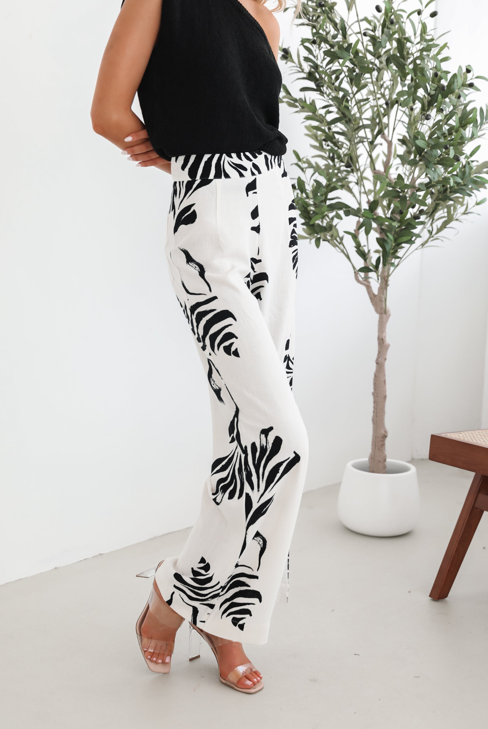 A woman sitting on a wooden chair wearing a black off-the-shoulder top paired with high-waisted white wide-leg pants featuring bold black tropical leaf prints. She completes the look with nude heels and is posed in a minimalistic room with white curtains and a potted plant in the background.