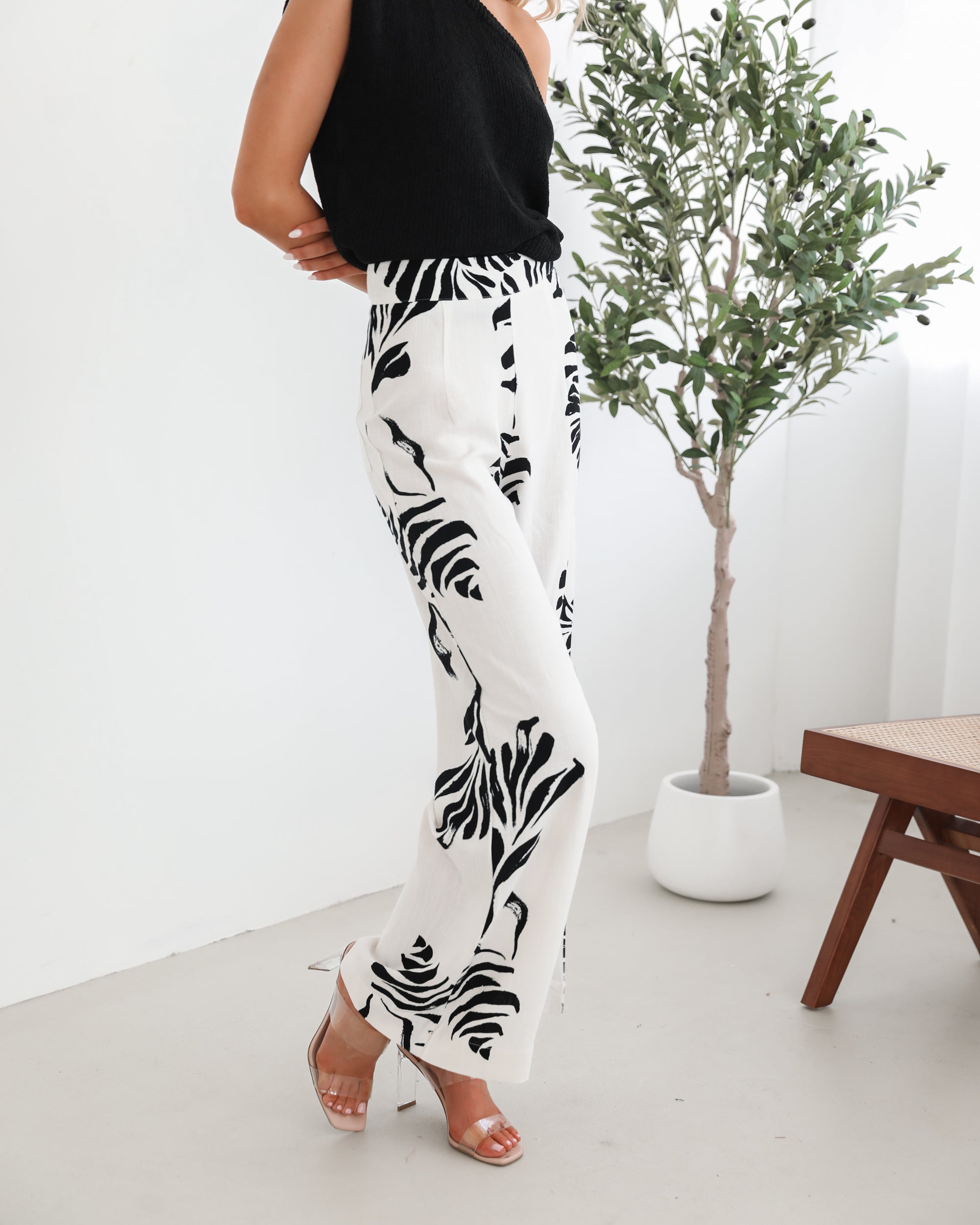 A woman sitting on a wooden chair wearing a black off-the-shoulder top paired with high-waisted white wide-leg pants featuring bold black tropical leaf prints. She completes the look with nude heels and is posed in a minimalistic room with white curtains and a potted plant in the background.
