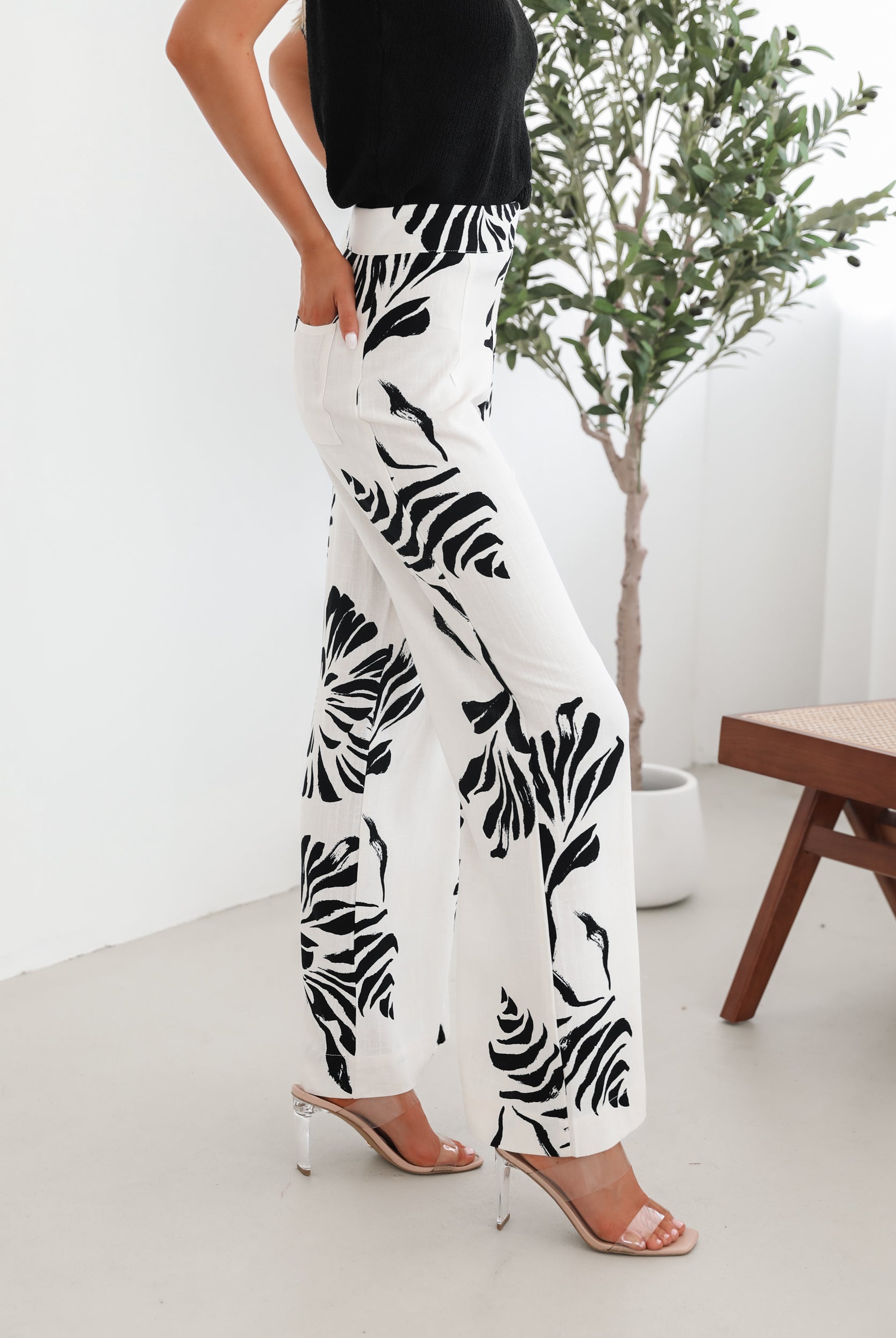 A woman sitting on a wooden chair wearing a black off-the-shoulder top paired with high-waisted white wide-leg pants featuring bold black tropical leaf prints. She completes the look with nude heels and is posed in a minimalistic room with white curtains and a potted plant in the background.