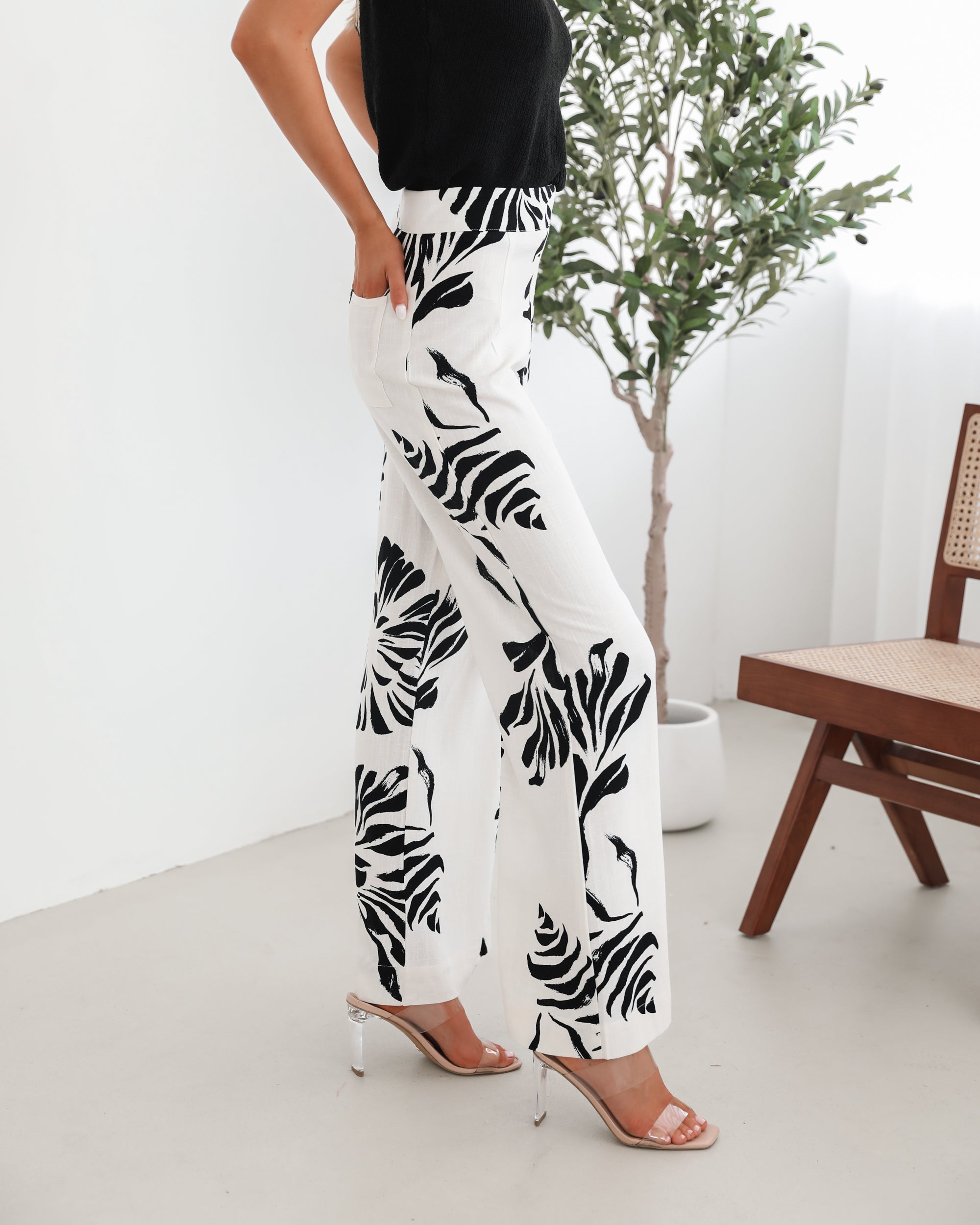 A woman sitting on a wooden chair wearing a black off-the-shoulder top paired with high-waisted white wide-leg pants featuring bold black tropical leaf prints. She completes the look with nude heels and is posed in a minimalistic room with white curtains and a potted plant in the background.