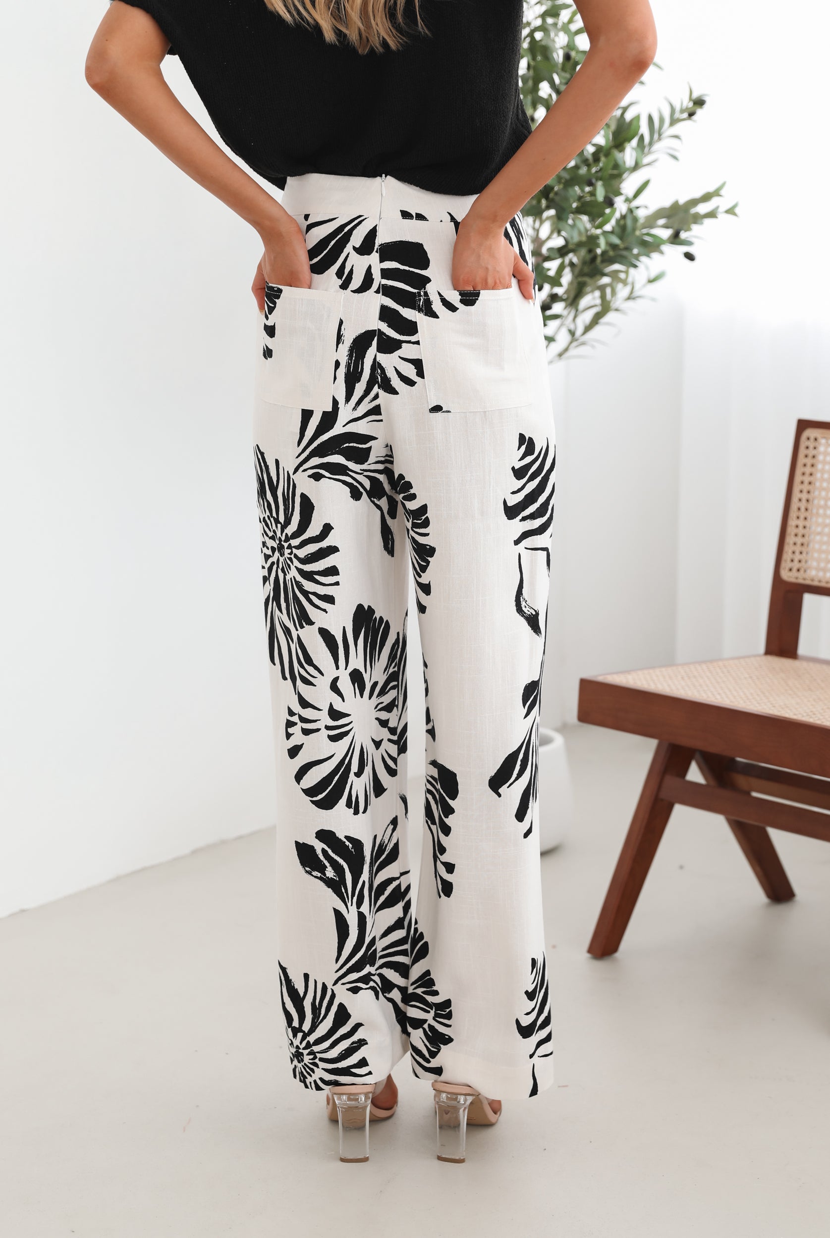 A woman sitting on a wooden chair wearing a black off-the-shoulder top paired with high-waisted white wide-leg pants featuring bold black tropical leaf prints. She completes the look with nude heels and is posed in a minimalistic room with white curtains and a potted plant in the background.