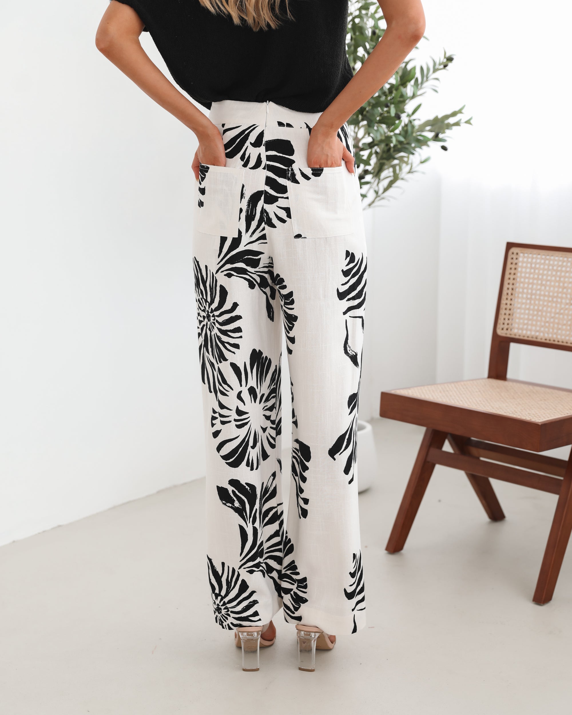 A woman sitting on a wooden chair wearing a black off-the-shoulder top paired with high-waisted white wide-leg pants featuring bold black tropical leaf prints. She completes the look with nude heels and is posed in a minimalistic room with white curtains and a potted plant in the background.