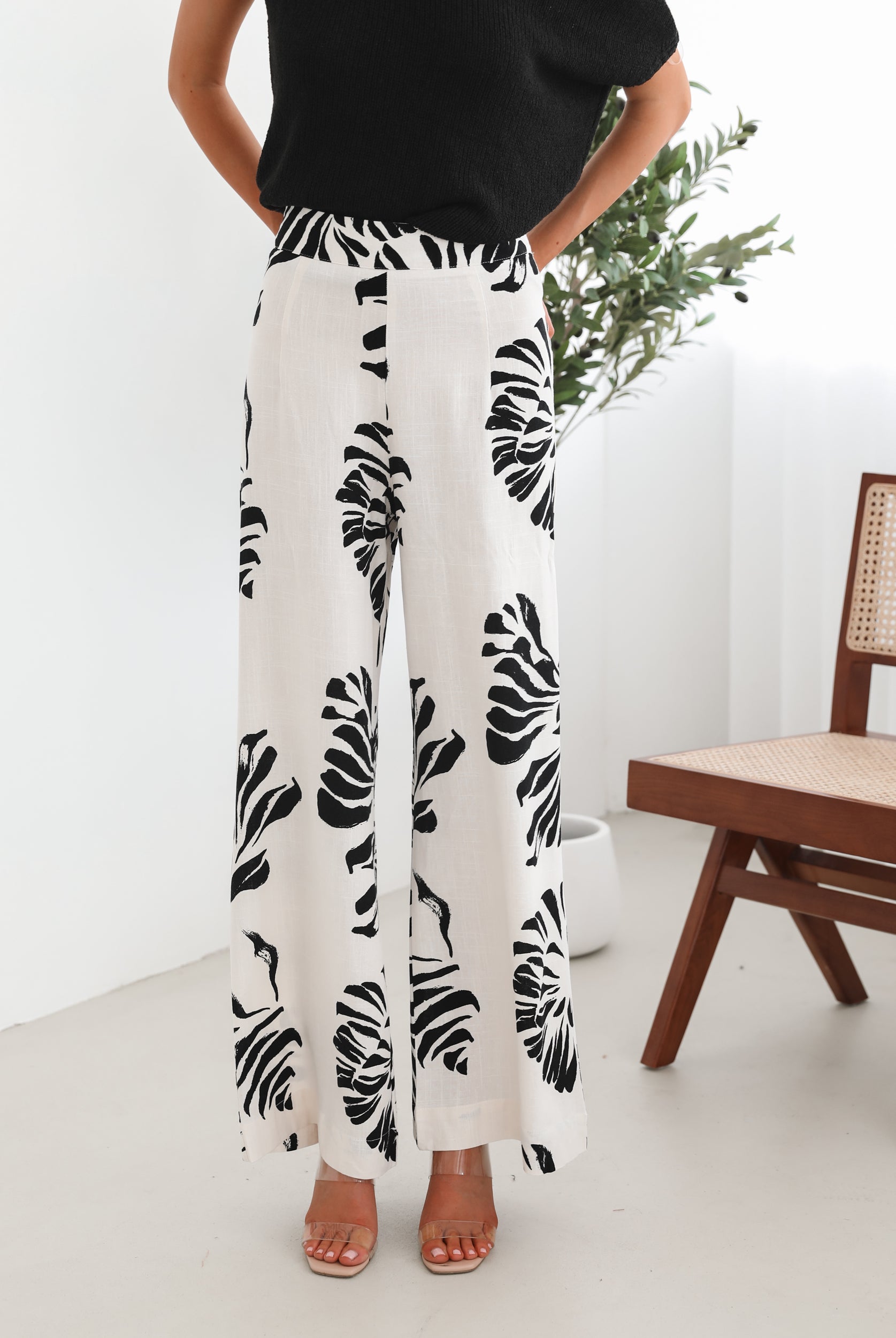 A woman sitting on a wooden chair wearing a black off-the-shoulder top paired with high-waisted white wide-leg pants featuring bold black tropical leaf prints. She completes the look with nude heels and is posed in a minimalistic room with white curtains and a potted plant in the background.