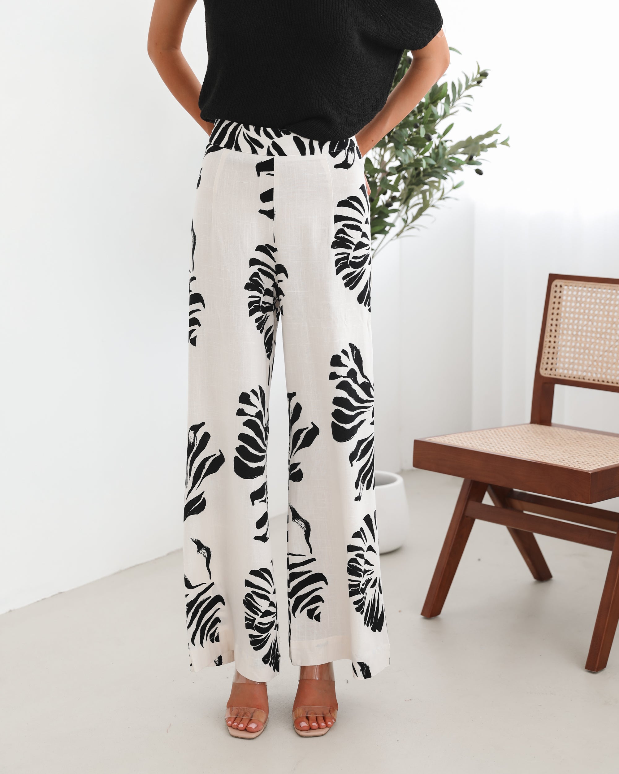 A woman sitting on a wooden chair wearing a black off-the-shoulder top paired with high-waisted white wide-leg pants featuring bold black tropical leaf prints. She completes the look with nude heels and is posed in a minimalistic room with white curtains and a potted plant in the background.
