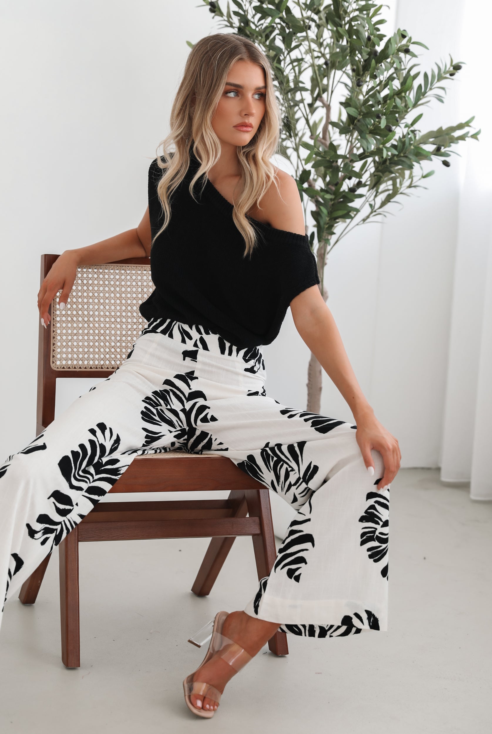 A woman sitting on a wooden chair wearing a black off-the-shoulder top paired with high-waisted white wide-leg pants featuring bold black tropical leaf prints. She completes the look with nude heels and is posed in a minimalistic room with white curtains and a potted plant in the background.