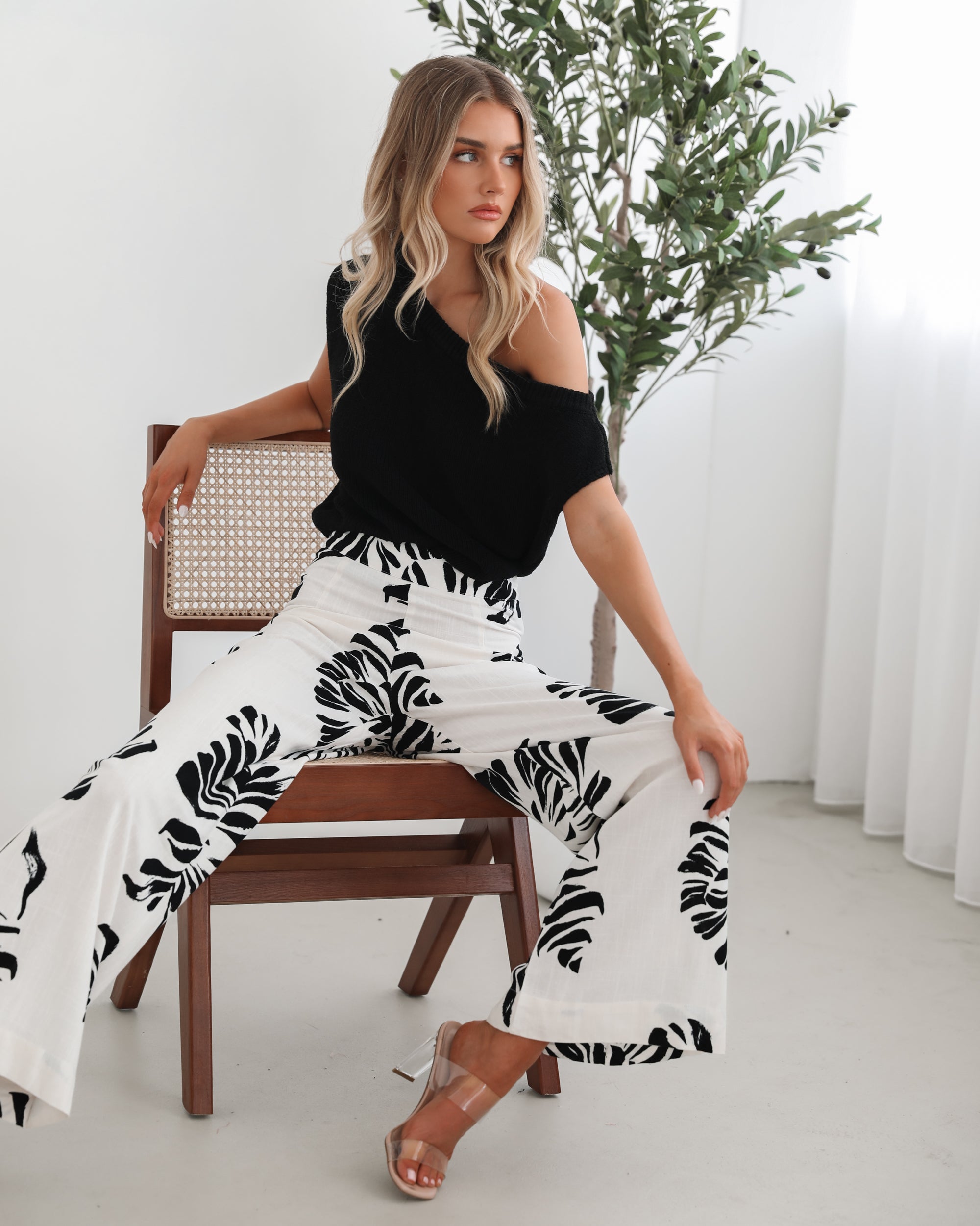 A woman sitting on a wooden chair wearing a black off-the-shoulder top paired with high-waisted white wide-leg pants featuring bold black tropical leaf prints. She completes the look with nude heels and is posed in a minimalistic room with white curtains and a potted plant in the background.