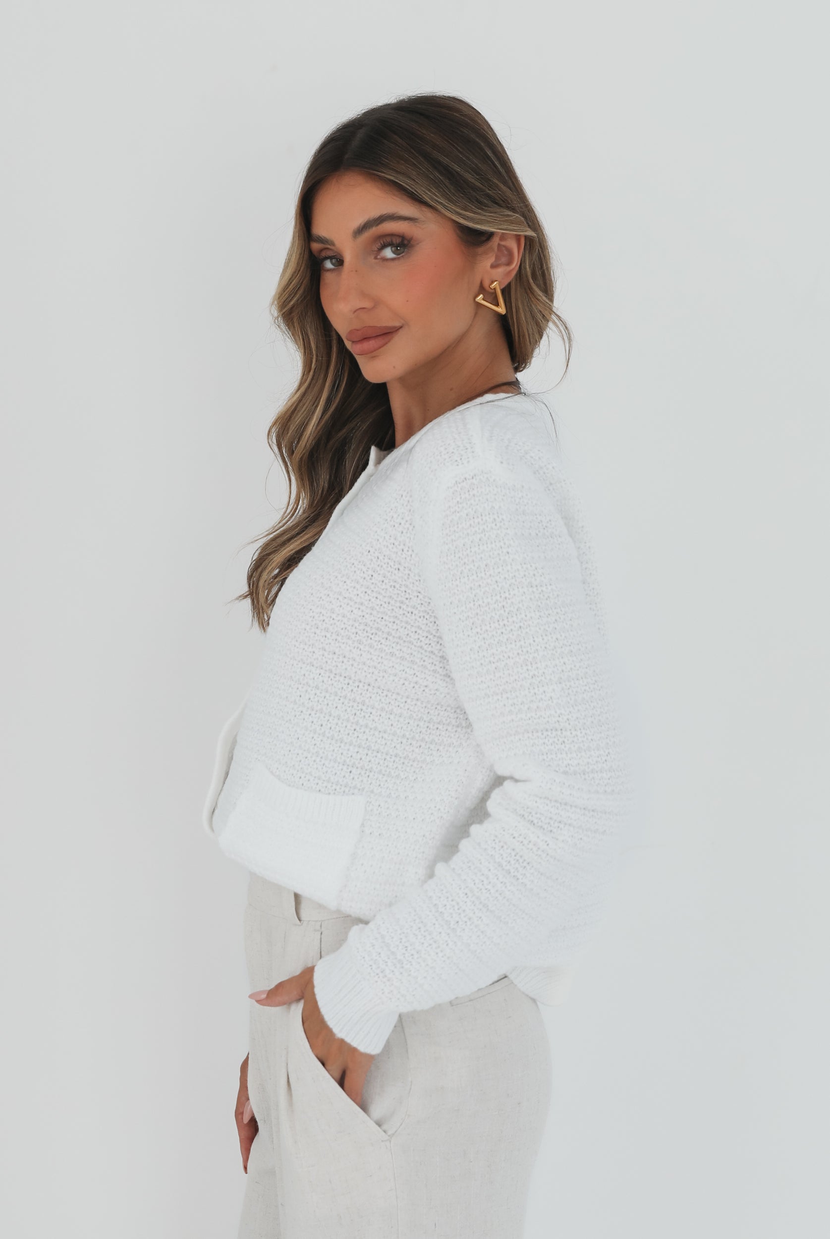 A woman wearing a white textured knit cardigan with front buttons and two small front pockets, paired with high-waisted light beige trousers. She stands against a plain white background, with one hand in her pocket and the other touching her hair.